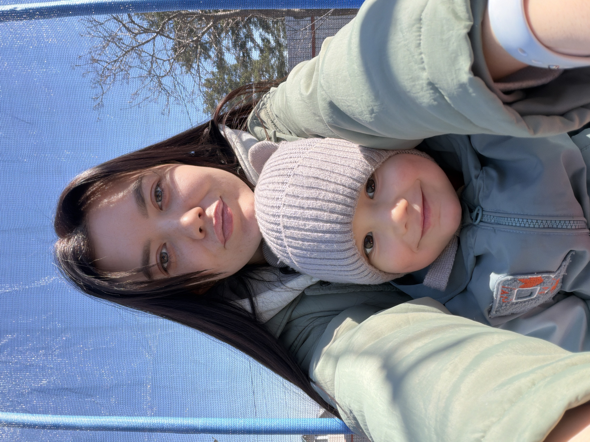 A medium-range, outdoor selfie captures a young woman and a small child, possibly on a trampoline. The woman has long, dark hair and light skin, and she is wearing a light green jacket. The child, also fair-skinned, wears a grey knitted hat. The background shows a blue, net-like material, likely the trampoline enclosure, and trees against a bright sky. The location is the city of Chaikovsky, Russia.