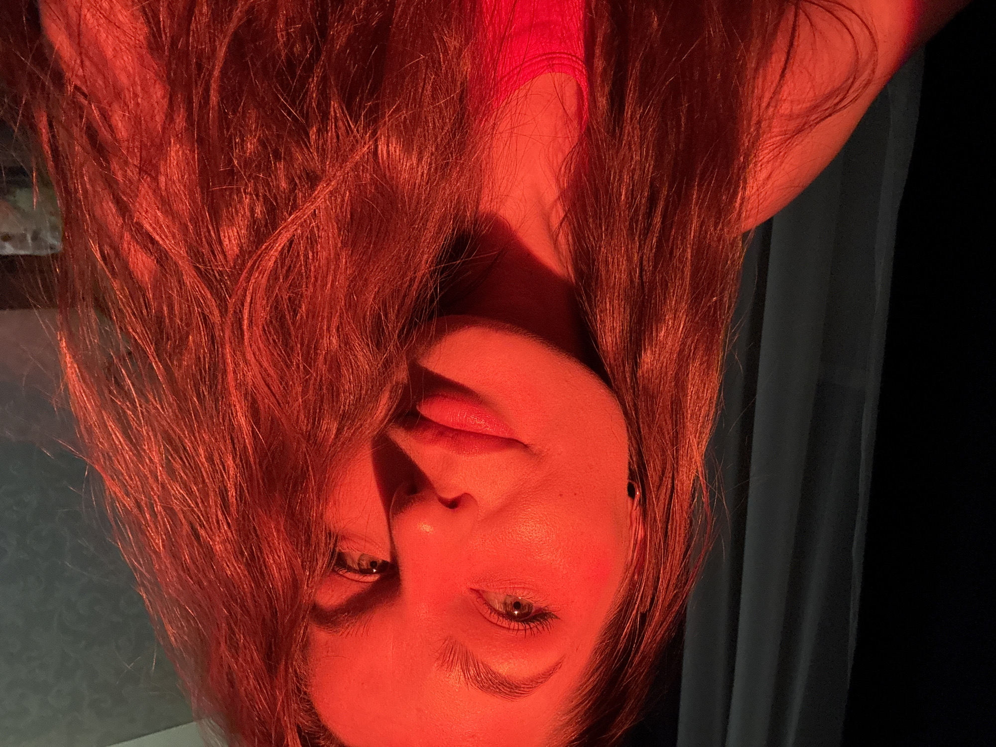 A portrait of a fair-skinned woman with long, auburn hair is illuminated with a red light. The woman is facing the camera, her head tilted so that her face appears upside down. Her eyes are open, and she has a neutral expression. The background consists of a dark curtain to the right and a partially visible textured wall to the left. The location is unknown, and there is no indication that the image was taken in Chaikovsky, Russia.