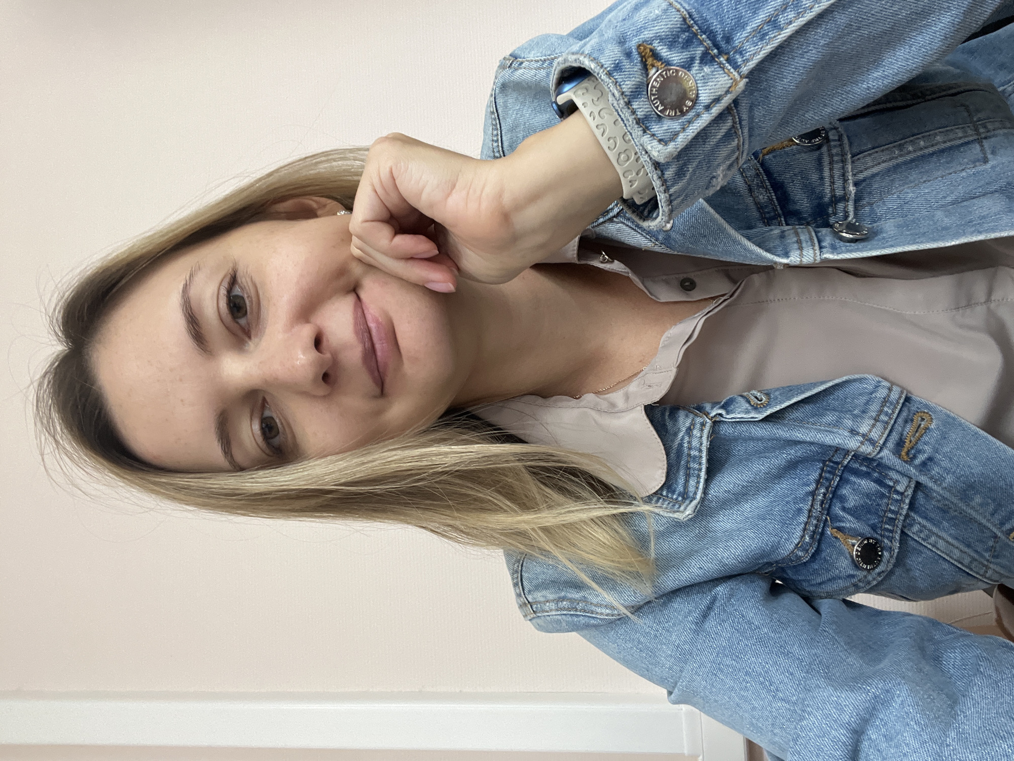 A mid-shot photograph shows a woman with shoulder-length blonde hair looking towards the camera and resting her chin on her hand. She is wearing a light-colored collared shirt under a blue denim jacket. Her right hand is visible, with fingers resting on her chin and cheek. She has a subtle smile. The background is a plain, light-colored wall with a vertical white trim piece on the left side. The lighting appears to be from the front and slightly to the side. No specific landmarks of Volgograd, Russia, are visible in this image.