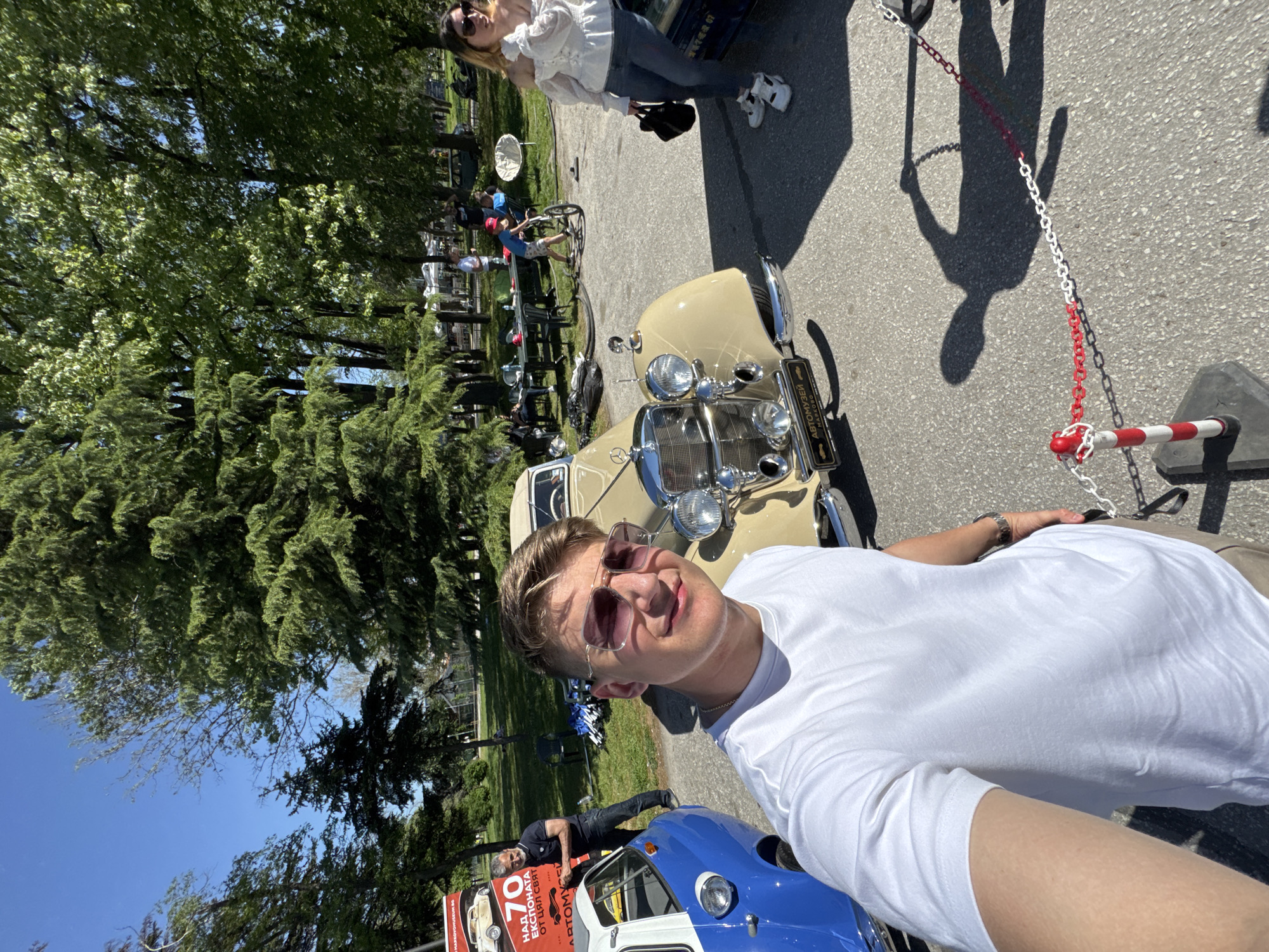 The image depicts a man taking a selfie at the "Retro Museum" in Plovdiv, Bulgaria. He is wearing sunglasses and a white t-shirt. Behind him are two vintage cars, one beige and one blue. In the background, there are other people, trees, and possibly part of the museum or exhibits. The area seems to be outdoors with paved ground. A woman stands near the beige car. A chained bollard is visible on the right. A poster ad is visible on the left. The sky is blue.