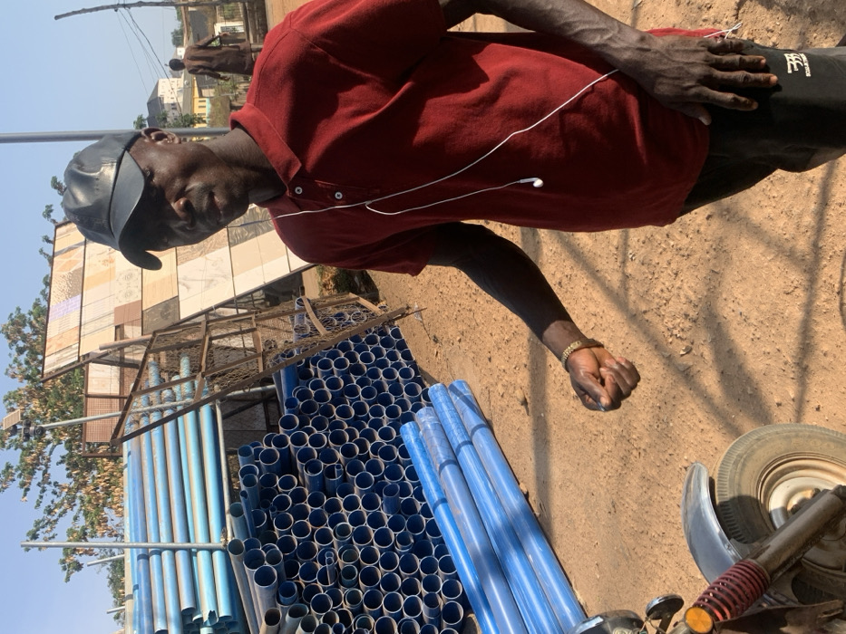 The image displays an outdoor scene in Magongo, Nigeria, under direct sunlight. In the foreground, an adult male with dark skin is positioned centrally, facing forward. He wears a dark baseball cap, a short-sleeved maroon polo shirt, and dark trousers. White wired earphones are visible against his shirt. A gold-colored bracelet adorns his left wrist, and his right hand is clenched. To the subject's left, large quantities of blue PVC pipes of varying diameters are stacked on the reddish-brown unpaved ground and on a multi-tiered metal rack. Behind these pipes, a tall metal frame displays numerous square tiles of diverse patterns and colors. In the distant background, a second male figure is partially visible, wearing light-colored clothing. A utility pole with what appears to be a street light or solar panel assembly stands further back, along with overhead utility lines. The bottom left quadrant shows the front wheel, fender, and suspension components of a motorcycle or a three-wheeled vehicle. Trees and faint outlines of buildings are visible in the far background.