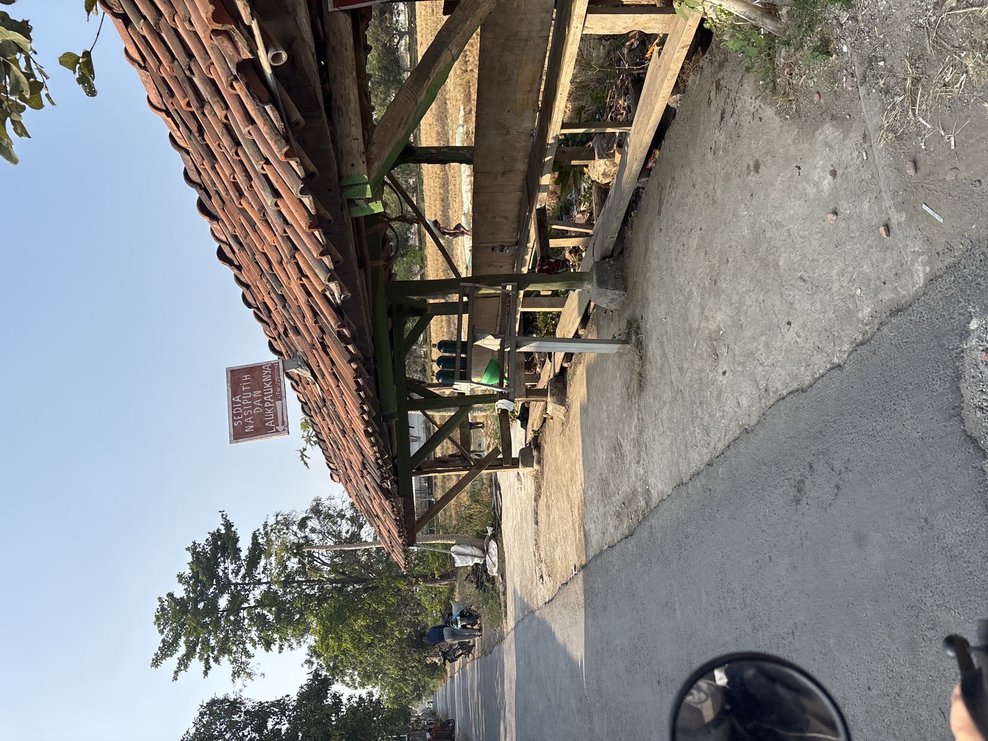 In Salawana, Indonesia, a roadside stall with a tiled roof is pictured. The stall is constructed with wooden beams and has an open-air design, with benches for seating. Several green gas canisters are visible within the stall. A sign is mounted on the roof, displaying text in red letters on a brown background, which reads "SEDIA NASI PUTIH DAN LAUK PAUKNYA," meaning "White rice and its side dishes are available." A paved road runs alongside the stall, with a few motorcycles parked near it. In the background, a person is walking by, and a field of dry crops is visible. The sky is clear and blue, indicating a sunny day.
