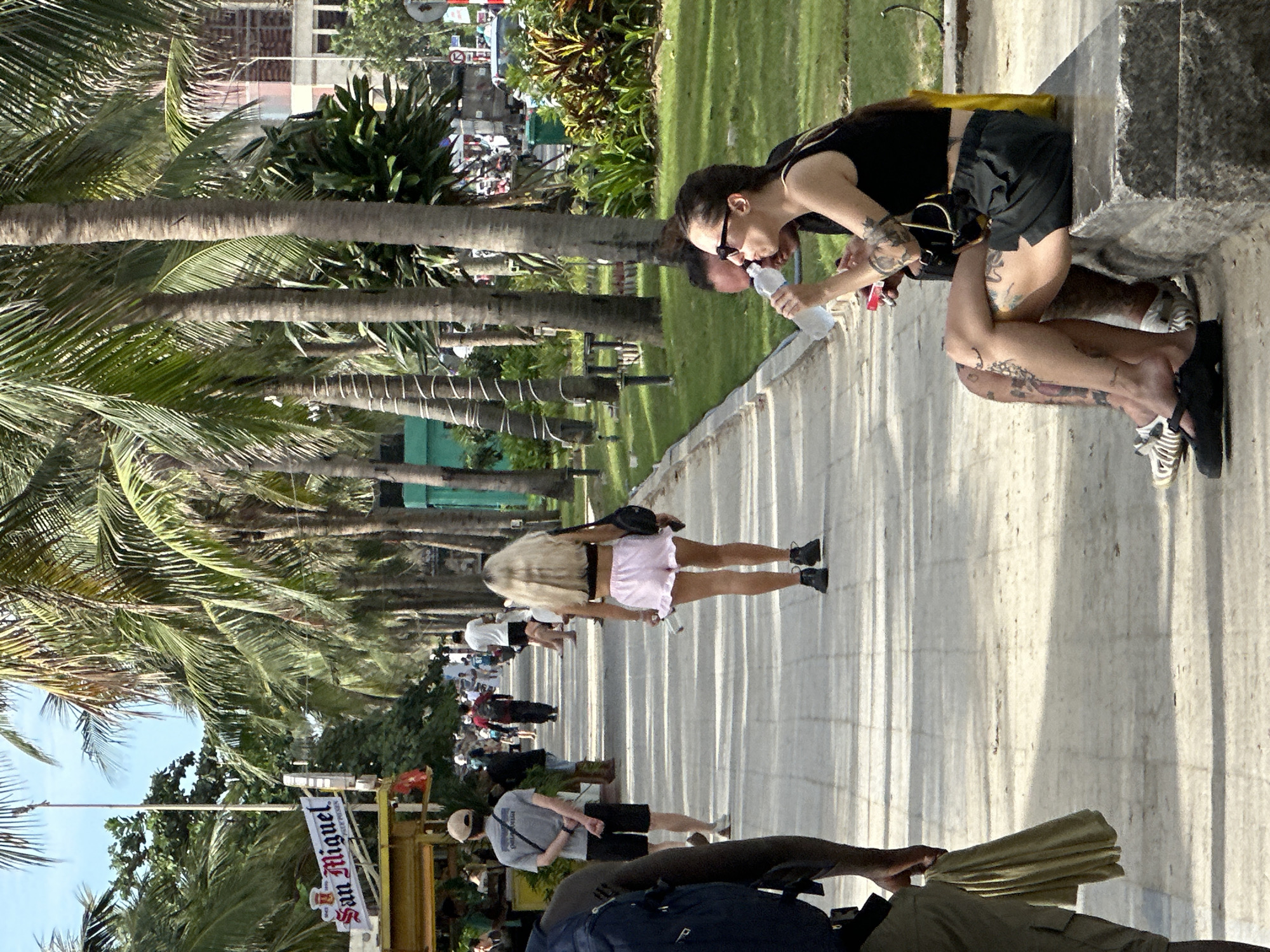 The image depicts a sunny daytime scene along a palm-lined promenade in Da Nang, Vietnam. In the foreground, a man with a backpack walks away from the camera, while a woman with blonde hair and pink ruffled shorts walks towards the camera. Further down the path, several other individuals are seen walking, and some are sitting on a stone bench. The woman on the bench is drinking from a water bottle. The environment includes a paved walkway, lush green grass, a row of tall palm trees, and some distant buildings. A food stall with a "San Miguel" sign is visible on the left side of the image. The overall atmosphere suggests a leisurely day in a tropical urban setting.