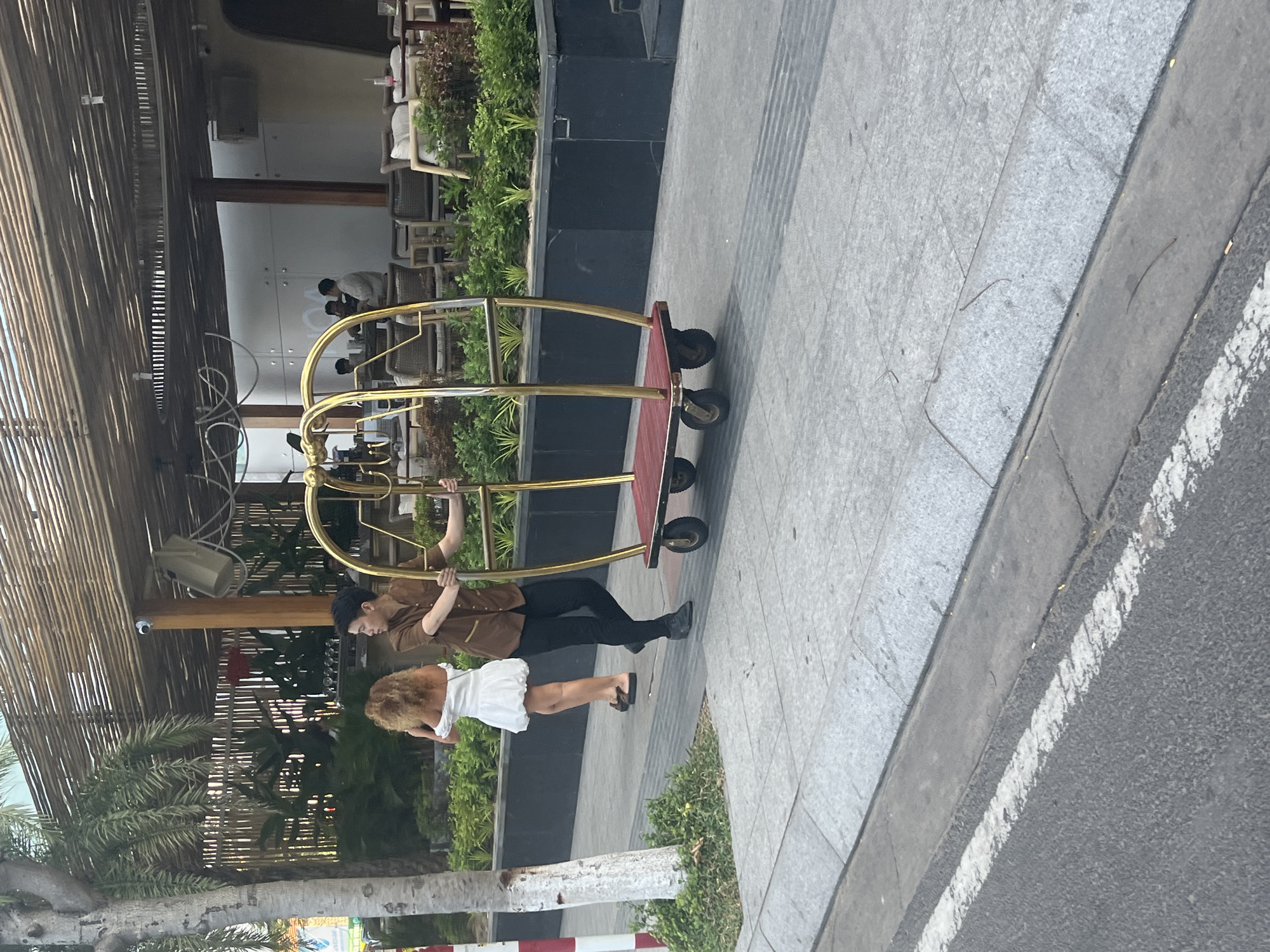 A hotel porter, dressed in a brown collared shirt and black pants, is pushing a golden luggage cart with a red base on a paved sidewalk in Nha Trang, Vietnam. A woman in a white off-the-shoulder dress walks away from the cart. Behind them, a palm tree and lush green plants surround a building with a bamboo-slatted roof, suggestive of an outdoor seating area for a restaurant or hotel. The scene appears to be on a sunny day, with shadows cast on the sidewalk.