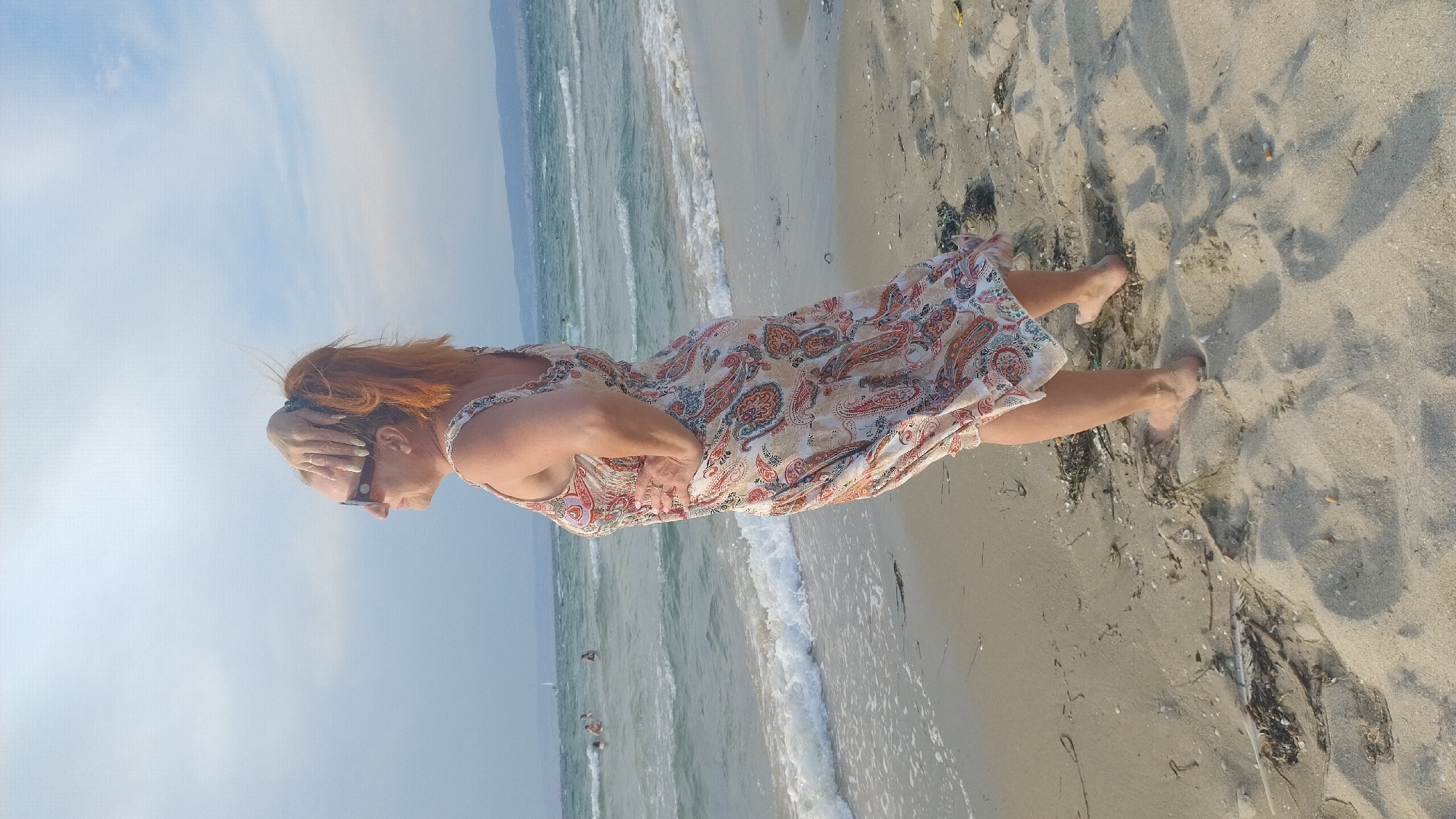 An adult female with reddish-orange hair tied in a ponytail, wearing sunglasses and a patterned sleeveless dress, stands on a sandy beach in Neoi Epivates, Greece. Her right hand is placed on top of her head, and her left hand is on her hip. She is looking to her left, out towards the sea. The sea has moderate waves, and there are a few people swimming or wading in the distance. The sky is partly cloudy. The beach is composed of sand and small debris, with some seaweed visible.