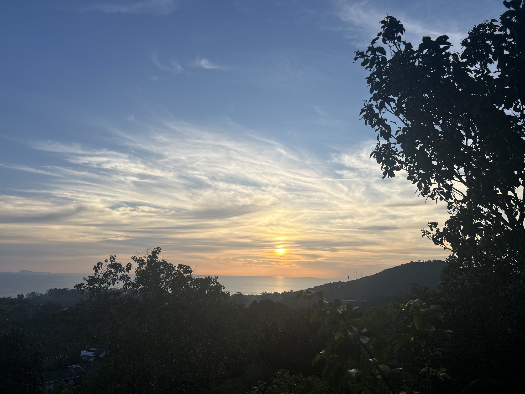 The image depicts a sunset over the sea in ตำบลเกาะพะงัน, Thailand. The sky is a gradient of blue to orange, with cirrus clouds spread throughout. The sun is a bright orb on the horizon, reflecting on the water. The foreground consists of silhouettes of trees and hills, suggesting a viewpoint from an elevated position. There is little to no activity, focusing instead on the natural beauty of the location.