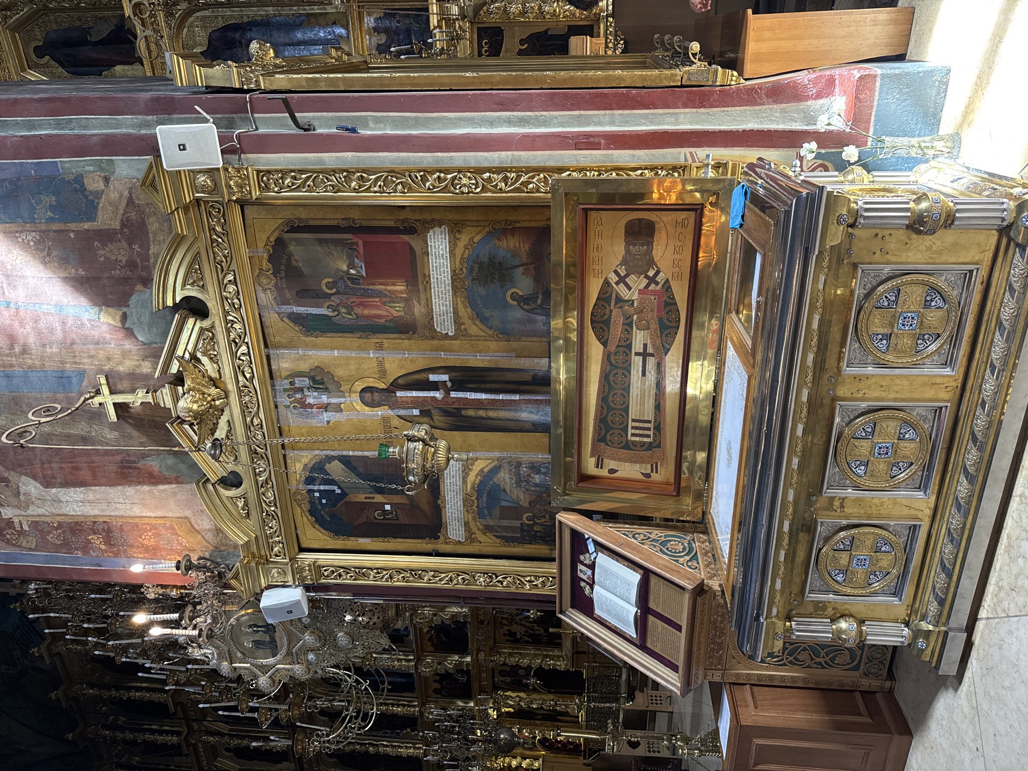 The image depicts the interior of the Dormition Cathedral within the Trinity Lavra of Saint Sergius in Sergiev Posad, Russia. The scene focuses on an ornate iconostasis, a gilded wooden screen with icons, which is a central feature of Orthodox churches. In the foreground, a richly decorated, gilded reliquary chest is prominently displayed, adorned with intricate patterns and medallions. To the left of the reliquary, a book is open on a stand, likely a holy text. Above the reliquary, a large icon is visible, depicting religious figures. The background reveals more of the cathedral's interior, including a large, multi-tiered chandelier with numerous candles, murals on the walls, and architectural details. The lighting suggests natural light streaming in from a window, illuminating parts of the scene. There are no people clearly visible in the image, but the presence of a blue cloth on the reliquary might indicate recent activity.