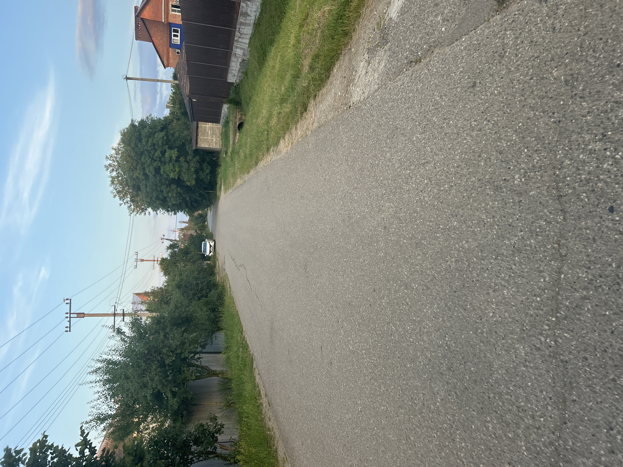 This is a wide-angle, low-angle shot of a paved road curving to the left, with a clear sky and some wispy clouds above. The image is oriented vertically. On the right side of the road, there is a grassy verge. Beyond the verge, on the left, are several elements of a rural or suburban setting.
In the foreground and midground, there is a dark brown, shed-like structure adjacent to a building with a reddish-brown brick facade. The building has a dark roof and several windows. A large, leafy green tree stands between the road and the buildings. Further down the road, more trees and vegetation line the left side, behind a grey metal fence. Power lines and utility poles are prominent in the background, stretching across the sky. A white car is visible on the road in the distance.
The lighting suggests it is daytime, likely late afternoon or early evening, given the soft light and the presence of clouds. The overall scene depicts a quiet, rural road in what appears to be a residential area. There are no people visible in the image. The mood is peaceful and serene. No text is visible in the image.