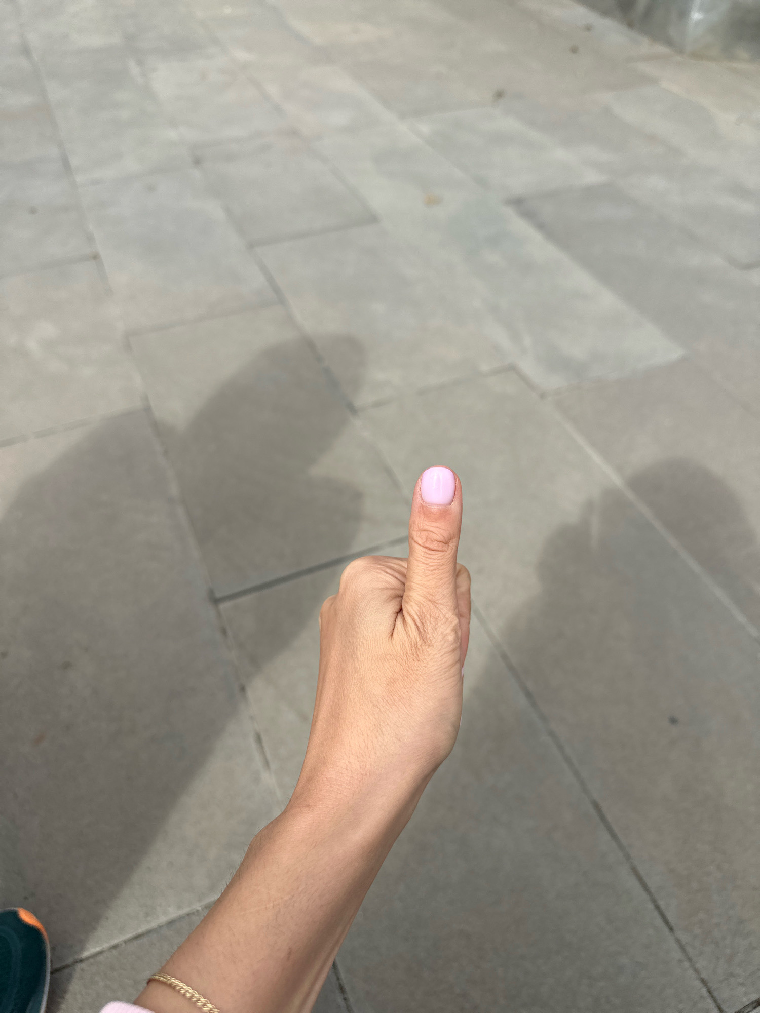 A hand with light skin, painted with light pink nail polish, is shown giving a thumbs-up gesture against a backdrop of grey paving stones. The paving stones are arranged in a rectangular pattern. Shadows of what appear to be a person and possibly other objects are cast on the ground, suggesting an outdoor setting. A hint of a green and orange athletic shoe is visible in the lower left corner of the frame, indicating the presence of at least one person. The scene does not provide any specific indicators for its location in Gyumri, Armenia.