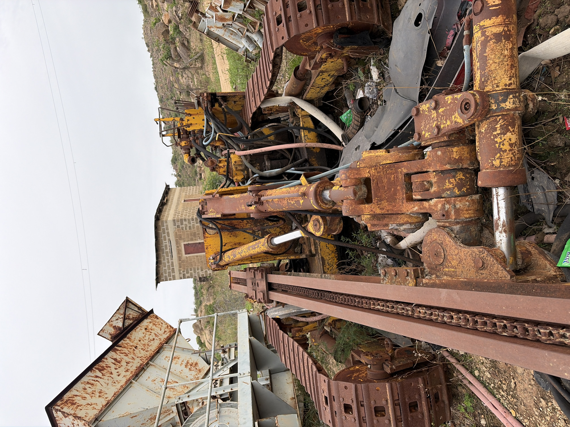 The image depicts a derelict industrial site in Gura Top, Nigeria. The focus is on a dismantled, rusting yellow-painted tracked vehicle, possibly a drilling rig or bulldozer, amid scattered metal debris and overgrown vegetation. The vehicle's hydraulic cylinders, hoses, and track components are prominently displayed. A building constructed of grey cement blocks is partially visible in the background. The environment appears to be an outdoor storage or salvage yard, with rocky hills visible in the distance under a cloudy sky. There are no people visible in the image.