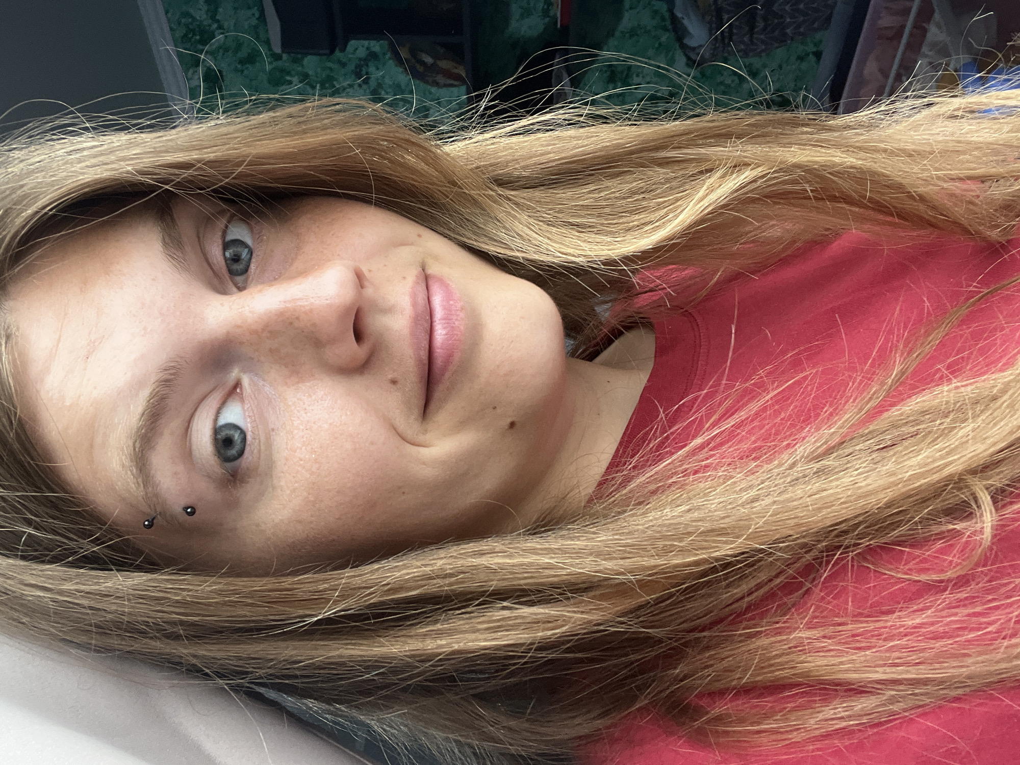 A medium shot shows a young woman with long, blonde hair and blue eyes looking slightly off-camera to her right. She has a faint smile and several freckles across her nose and cheeks. She is wearing a bright red t-shirt. An eyebrow piercing is visible on her left eyebrow. The background is softly blurred, showing hints of green textured wallpaper and some shelving with objects. The image appears to be taken indoors. The provided location of Сеніцкі сельскі Савет, Belarus, is not discernible from the image itself.