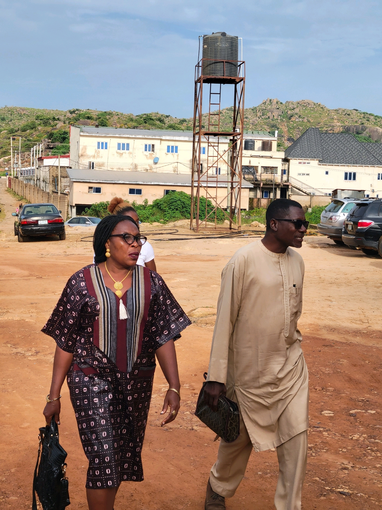 In Gura Top, Nigeria, two individuals, a woman and a man, walk outdoors on a dirt path. The woman, in the foreground, wears a patterned dress and glasses. She carries a black purse. The man, to her right, is dressed in a light-colored tunic and pants and wears sunglasses. He holds a patterned bag. In the background, a water tower stands tall, and several buildings and parked cars are visible. Rocky, green hills form the backdrop under a cloudy sky.