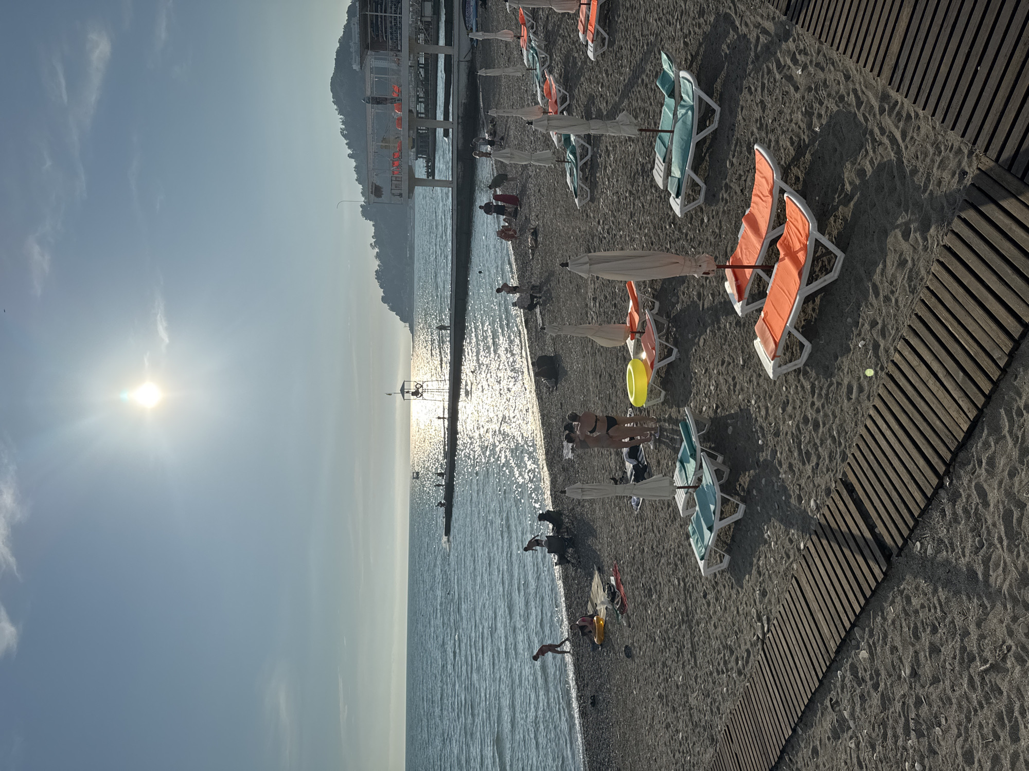 This is a photograph of a beach in Sochi, Russia, under a clear sky with the sun shining brightly. The beach is covered with dark sand and pebbles, with several wooden pathways leading to the water. Numerous lounge chairs, some with umbrellas, are set up on the sand. In the foreground, a wooden pathway is visible, and further back, lounge chairs with orange and turquoise cushions are scattered across the beach. People are seen scattered across the beach, some sunbathing, others walking near the water's edge. The sea is calm with gentle waves lapping the shore, and sunlight reflects off its surface, creating a shimmering effect. A pier extends into the sea, with a few figures visible on it. In the background, there is a building structure elevated on stilts, likely a cafe or restaurant, and beyond that, a forested hill. A lifeguard tower is also visible near the pier.