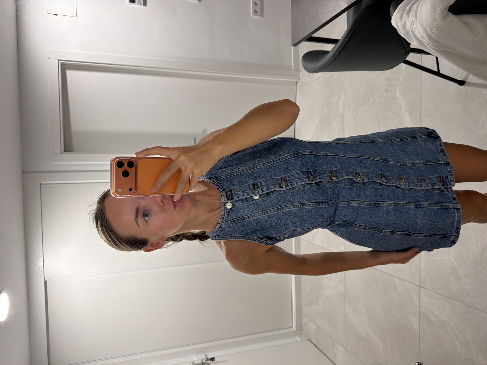 A woman is standing in a room and taking a mirror selfie. She is wearing a denim dress and holding an orange phone. The room appears to be a dressing room or bedroom. There are closets and a door in the background. A chair and a table are visible on the right side of the image. The floor is tiled. The overall lighting is bright. The image was taken in Finestrat, Spain.