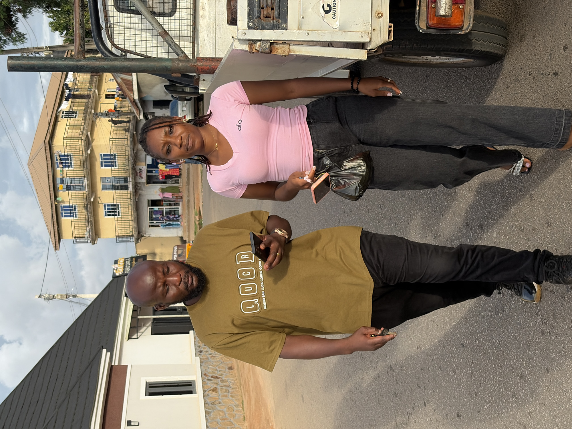 The image depicts a street scene in Jos, Nigeria, featuring two individuals walking on an asphalt road. A male subject is on the left, and a female subject is on the right, with various buildings and a utility truck in the background.
The male, an adult with a bald head and a dark beard, is wearing an olive-green t-shirt with "LUCR" and "BEHIND BAD LUCK COMES GOOD LUCK" printed in white on the front. He is wearing black trousers and dark sneakers. A gold-colored watch is on his right wrist, and he holds a black smartphone in his right hand, looking down at the screen. His left hand appears to hold keys or a similar small object.
The female, an adult with dark skin and hair styled in dreadlocks or braids, is wearing a pink V-neck t-shirt with the "alo" logo on the left chest. She wears dark grey, loose-fitting jeans and open-toed sandals with metallic embellishments. A gold-colored necklace with a small pendant is visible, along with a black beaded bracelet on her left wrist and a ring on her right hand. She holds a pink smartphone in her right hand, which also clutches a small black plastic bag. She is looking directly towards the camera with a neutral expression.
In the immediate background to the right of the subjects, a white utility truck is partially visible. It has a flatbed, a rear tire, a red taillight, and rusted metal components. A "CAUTION" sticker is present on its side.
The broader background features several buildings. To the far left, a modern-style house with a dark, pitched roof, light-colored walls, and stone cladding at the base is visible, along with a dark-framed window and an exterior light fixture. Towards the center-right, a prominent three-story yellow building with a brown roof is visible. This building has multiple blue-framed windows, balconies with black railings, and clothing items drying on several balconies. The ground floor appears to house shops with various garments displayed, and a partial sign reading "FABRIC CARE" is visible. Between the yellow building and the white house, a smaller building with a reddish-brown corrugated roof is present. A utility pole with overhead wires stands further back. The sky is partly cloudy, and the lighting indicates either early morning or late afternoon. The road surface is dark asphalt with visible aggregate.