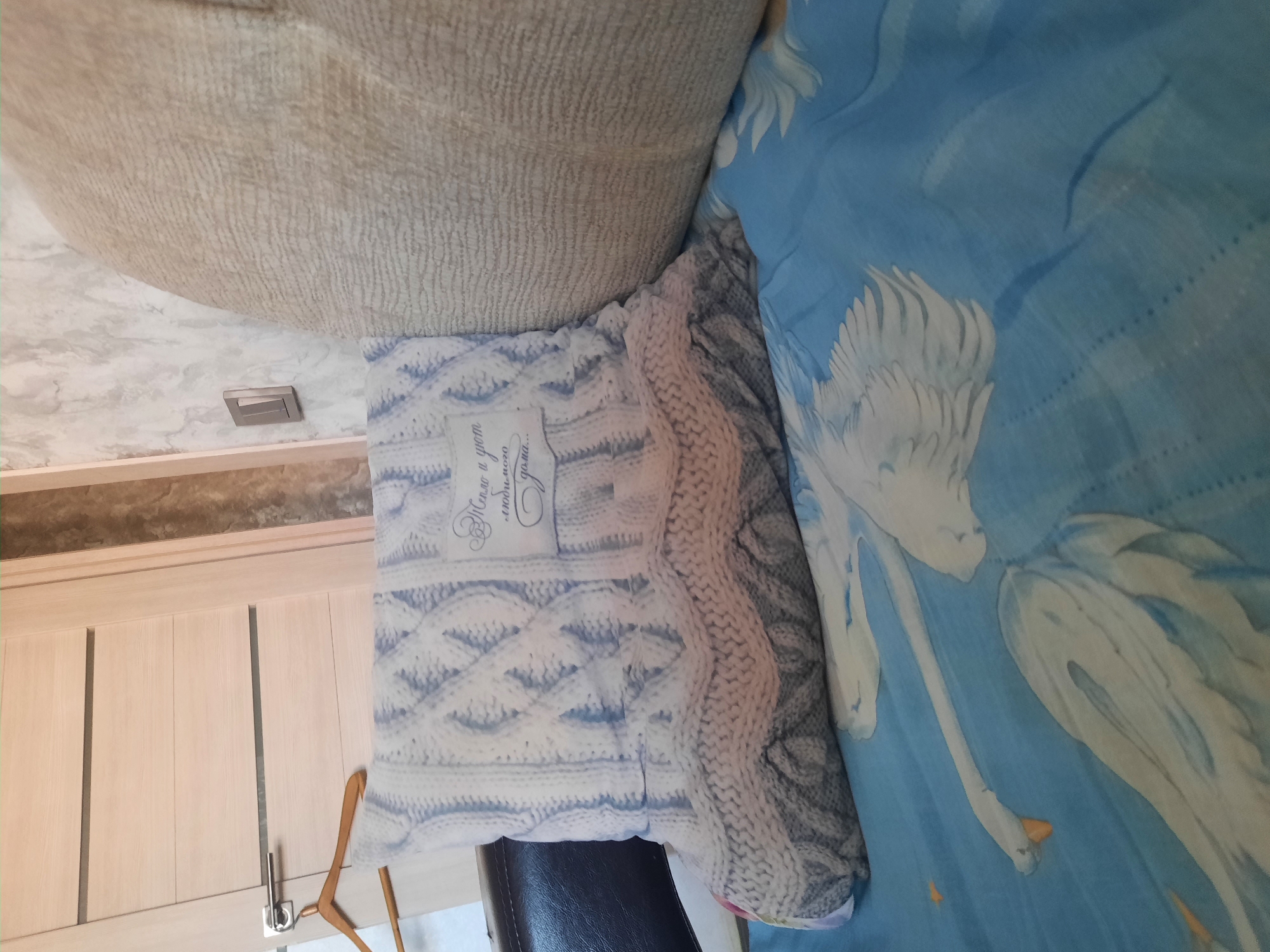 A soft focus medium shot shows a knitted pillow with a cable knit pattern in blue and white. A white label with Cyrillic text is attached to the front of the pillow. The pillow rests on a blue sheet with a swan motif. A light-colored upholstered armchair is visible to the right of the pillow. A wooden door with horizontal panels and a silver handle is in the background on the left. A wooden hanger hangs from the door handle.