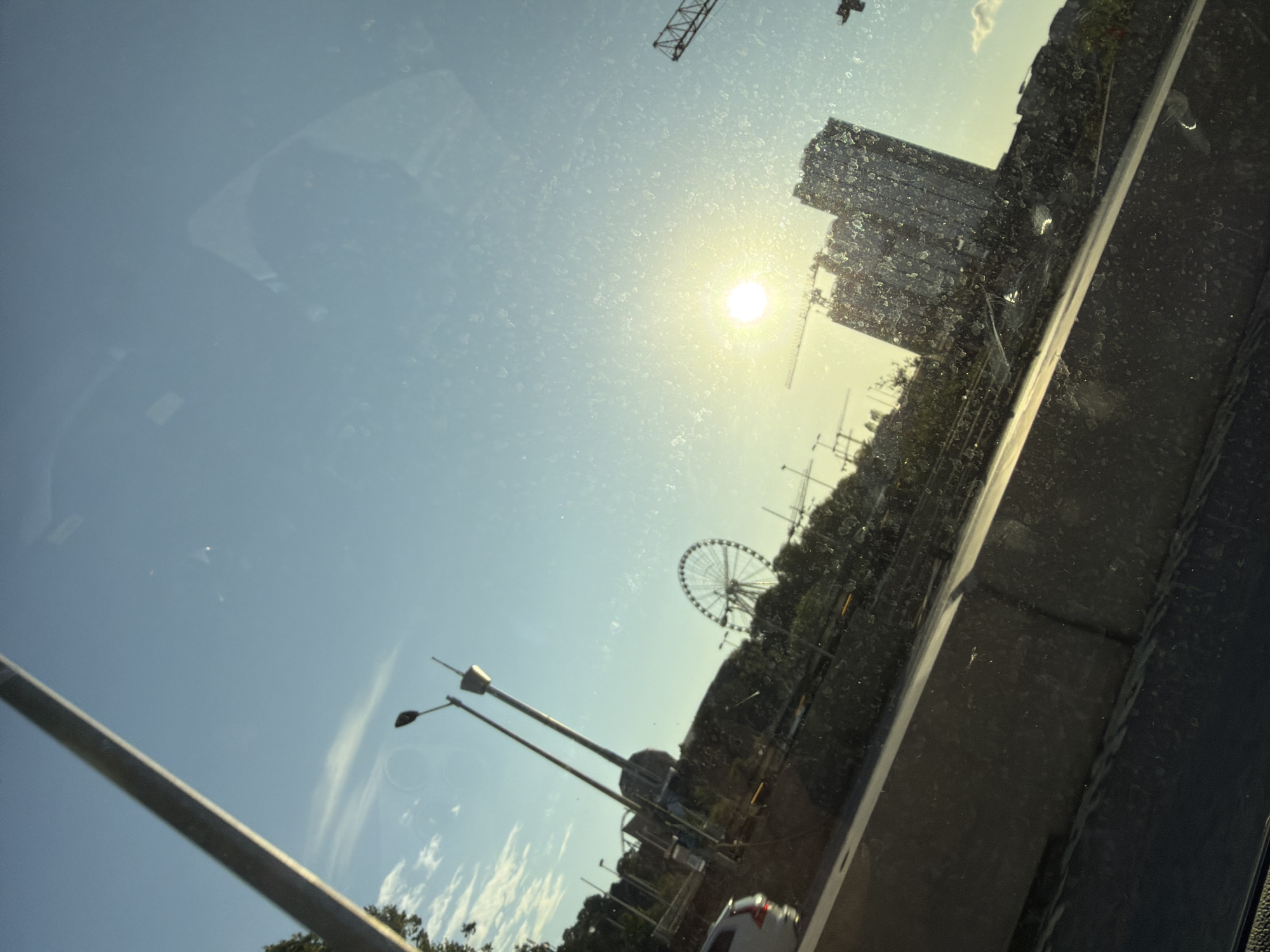 The image captures a sunlit cityscape, likely taken from within a vehicle, as suggested by reflections on the glass. In the distance, a prominent Ferris wheel stands against a clear blue sky with scattered white clouds. The sun, positioned in the upper right quadrant, casts a bright glare, partially obscuring details of several construction cranes and multi-story buildings. These structures appear to be in various stages of development, with scaffolding and unfinished elements visible on some. Trees and greenery line the midground, suggesting an urban park or landscaped area. A white car is partially visible in the lower left, hinting at the context of travel. The overall scene is bathed in the warm light of either sunrise or sunset, creating a tranquil yet dynamic urban environment. The distinctive architecture and presence of a Ferris wheel could indicate a location like Liseberg amusement park in Gothenburg, Sweden, known for its prominent Ferris wheel and its proximity to urban development and green spaces.