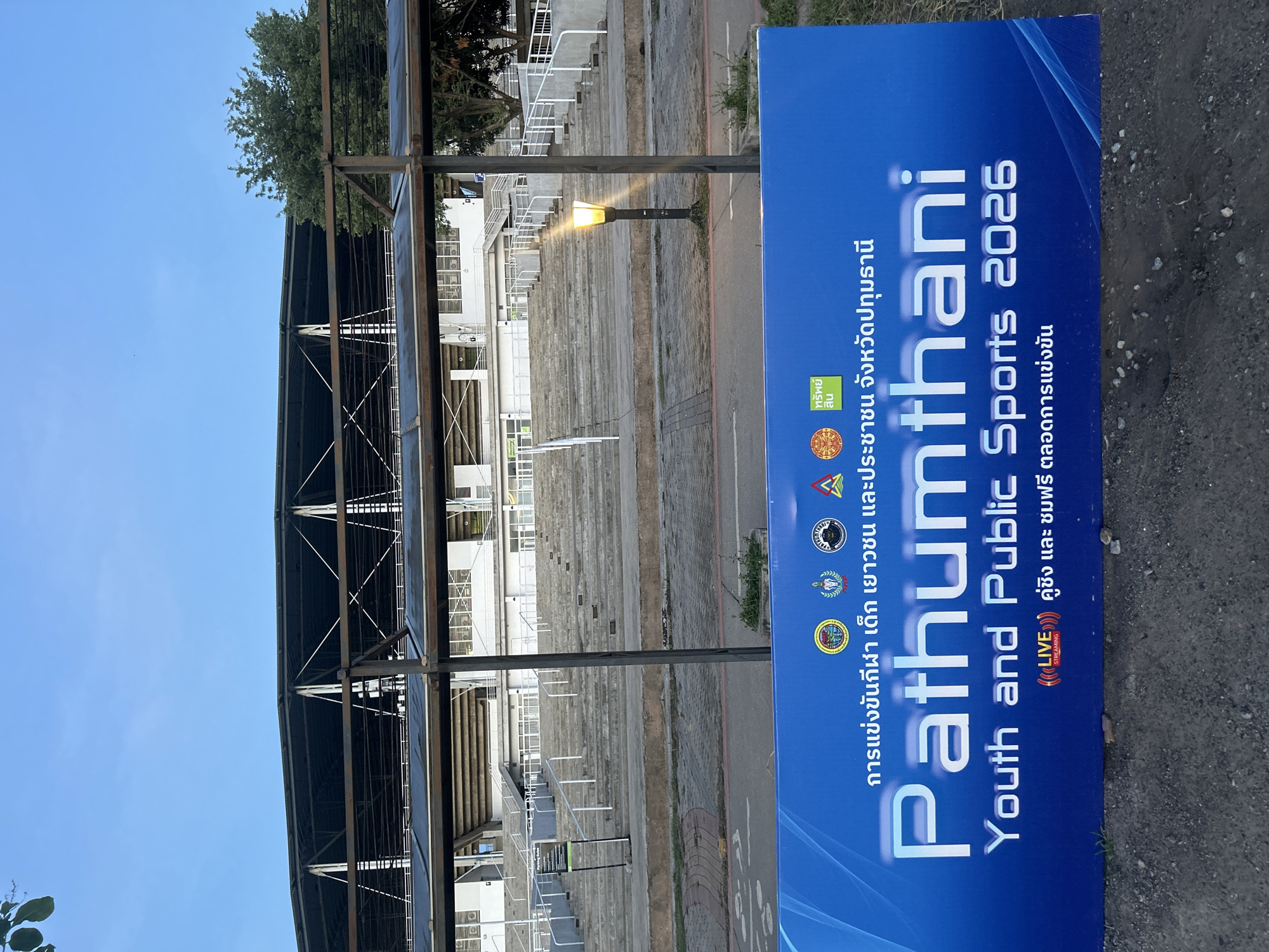 The image shows a stadium with a bright blue banner in the foreground. The banner has text in both Thai and English. The English text reads "Pathumthani Youth and Public Sports 2026". Below this, it says "LIVE STREAMING" with Thai text accompanying it. The stadium appears to be located in Tha Khlong Town Municipality, Thailand. The stadium has tiered seating, a covered roof supported by metal structures, and some trees visible in the background. The sky is a clear blue.