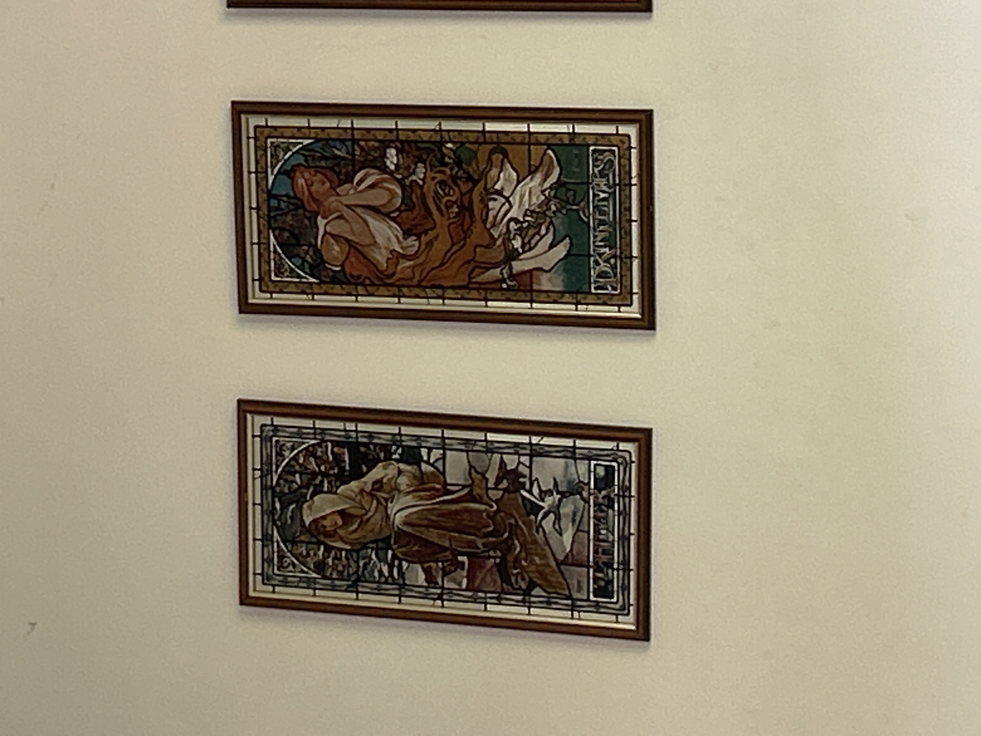 Two framed images are displayed on a light-colored wall. The frames are dark brown. The top image depicts figures in a stylized manner with a mix of brown, white, and green hues. A word is visible along the right edge, partially obscured. The bottom image shows similar figures, rendered in browns and grays against a dark background. The city and country are not discernible from the image.