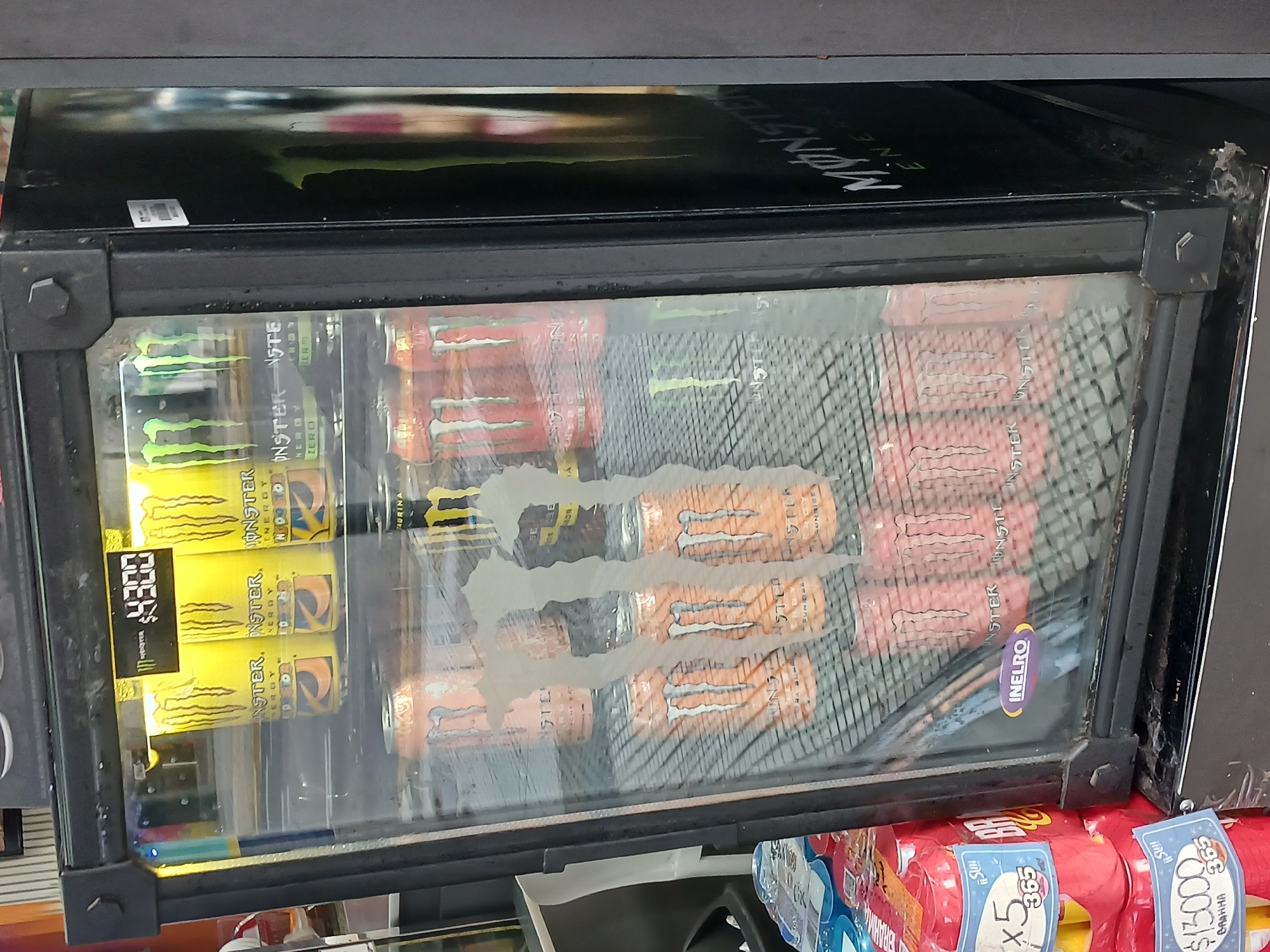 This image displays a refrigerated display cooler, likely found in a convenience store or kiosk in Buenos Aires, Argentina. The cooler is predominantly stocked with various flavors of Monster Energy drinks.
The main subjects are the Monster Energy cans. On the left, several green cans (likely Monster Energy Zero or Original) are visible, along with multiple yellow cans, which appear to be Monster Mango Loco or similar flavor, displaying a price tag of "$4300". To the right, numerous orange cans, possibly Monster Pipeline Punch or Pacific Punch, are stacked. Further right, red cans are partially visible. The cooler door shows condensation, indicating it is actively cooling the beverages.
The top of the cooler is branded with the "MONSTER ENERGY" logo. A small purple oval sticker with "NELRO" written on it is affixed to the glass door. Below the cooler, other packaged products are visible, including what appears to be packs of Brahma beer, with price tags such as "$13000" and another indicating "x5" and "$365".
Visible text from the cans includes "MONSTER ENERGY", "ZERO", "ENERGY", "TAURINA", and "ACHY BE". The prices on the tags, "$4300", "$13000", and "$365", refer to Argentine Pesos, consistent with the location context. The overall scene suggests a retail environment. There are no people visible, and the time of day and weather cannot be determined from the image.