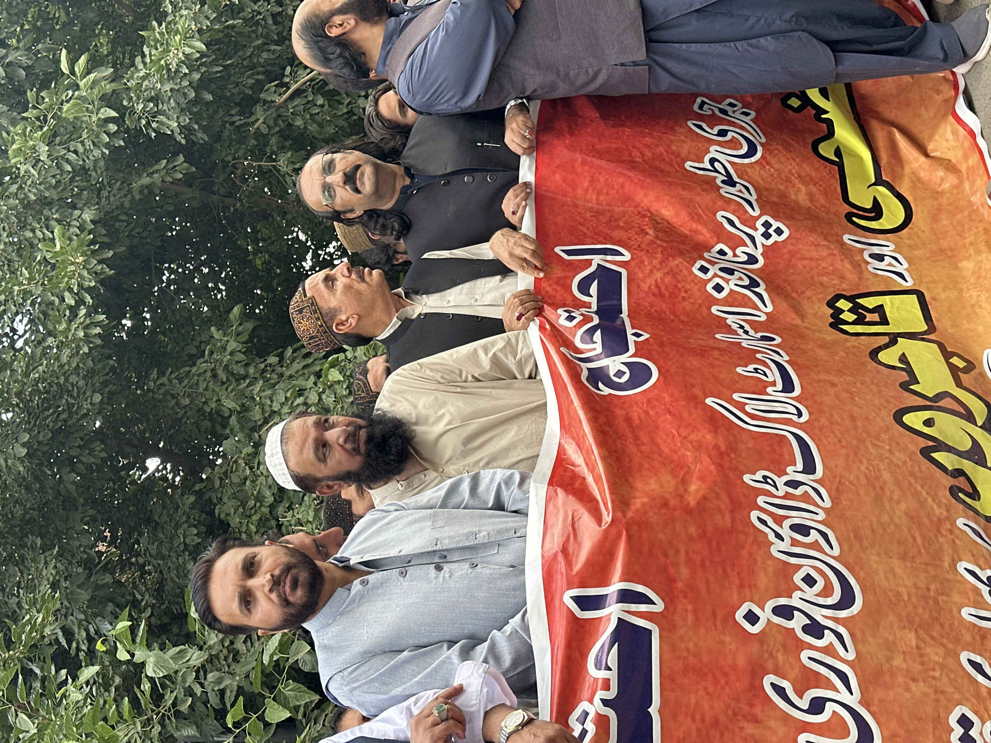 A group of men stand outside holding a red and orange banner with text written in Urdu or Arabic script. The men vary in age and are dressed in traditional Pakistani clothing such as shalwar kameez, vests, and skull caps. Some have beards and mustaches. The banner appears to be homemade and possibly related to a political or social cause. The background includes green trees and foliage, suggesting an outdoor location. The photo was taken in the city of Quetta City Tehsil, Pakistan.