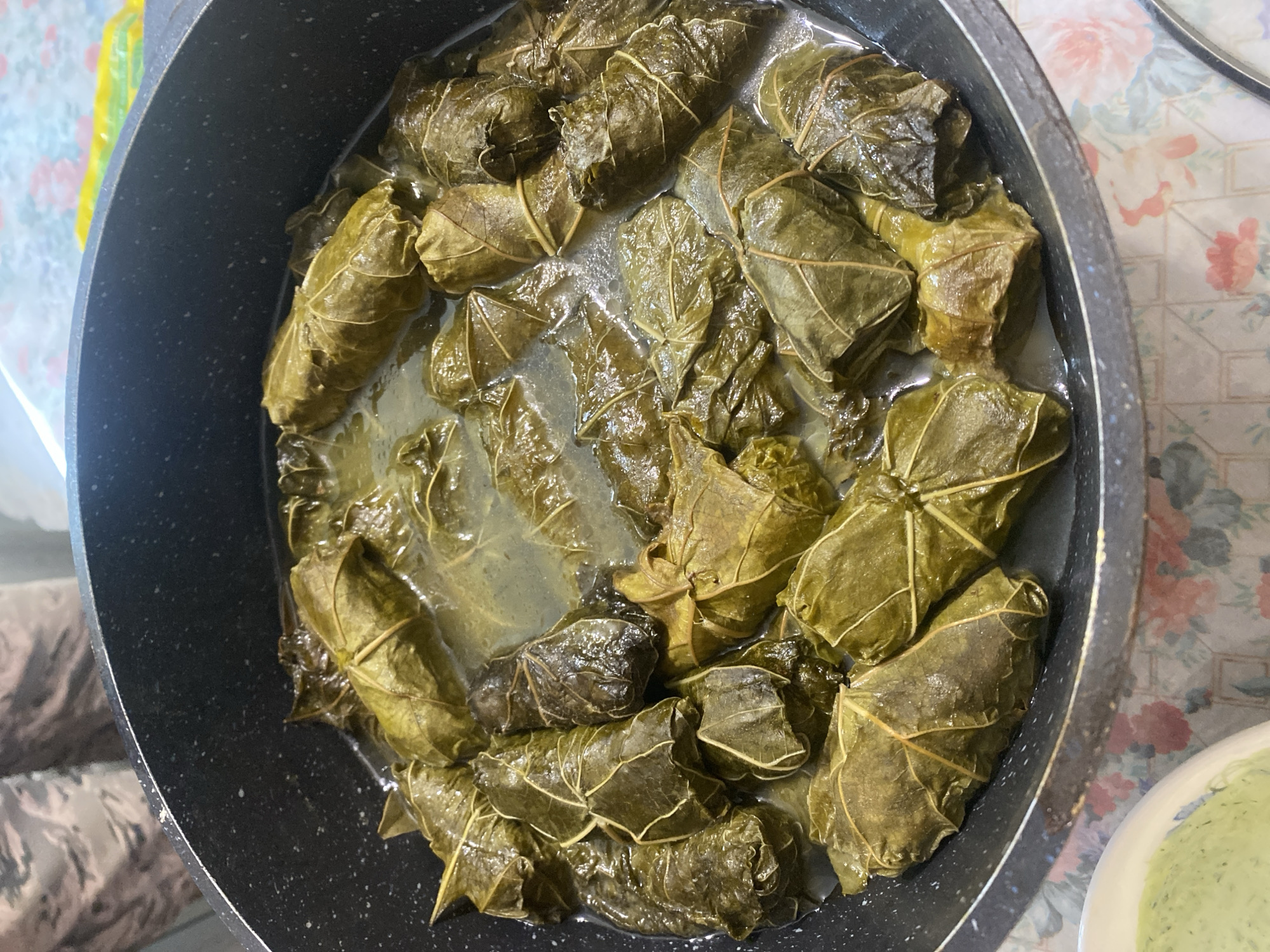 A top-down, close-up shot shows a pot of stuffed grape leaves, a dish known as dolma. The grape leaves are a deep green, some appearing wilted and darker, while others are a lighter, more vibrant green. They are tightly rolled and packed closely together in the pot, submerged in a light-colored liquid. The pot itself is black and appears to be made of a non-stick material, with a slightly textured interior. In the bottom left of the frame, a white bowl is partially visible, containing a pale green, creamy substance, likely a sauce or dip. The background is out of focus, but suggests a patterned tablecloth in shades of pink and white, and a person wearing camouflage-patterned pants. The image is well-lit, emphasizing the textures and colors of the food. This scene is not specific to Shiryaevo, Russia, and no elements within the image suggest this location.