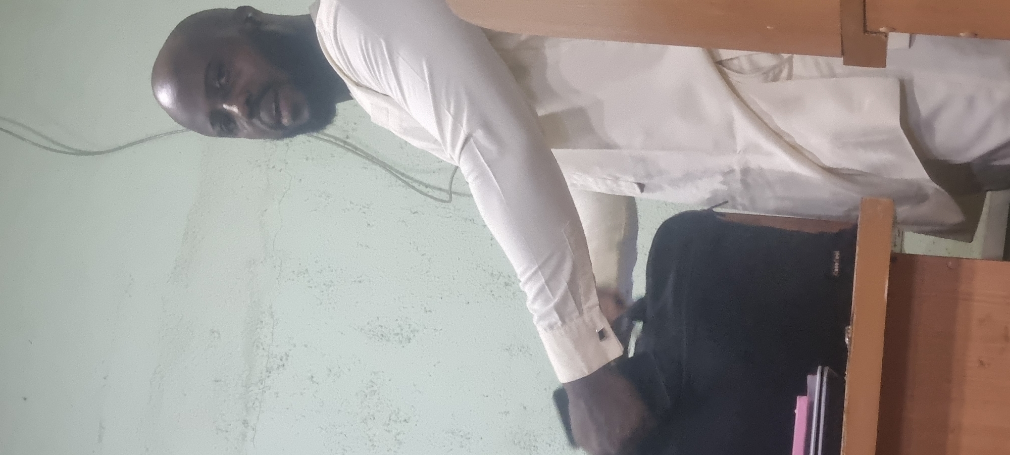 An exterior shot shows a man wearing a light-colored garment and black pants, sitting at a desk in Ikeja, Nigeria. He has a beard and is balding. He's looking to his left and speaking. A black backpack is on the desk, and a pink object is on top of it.