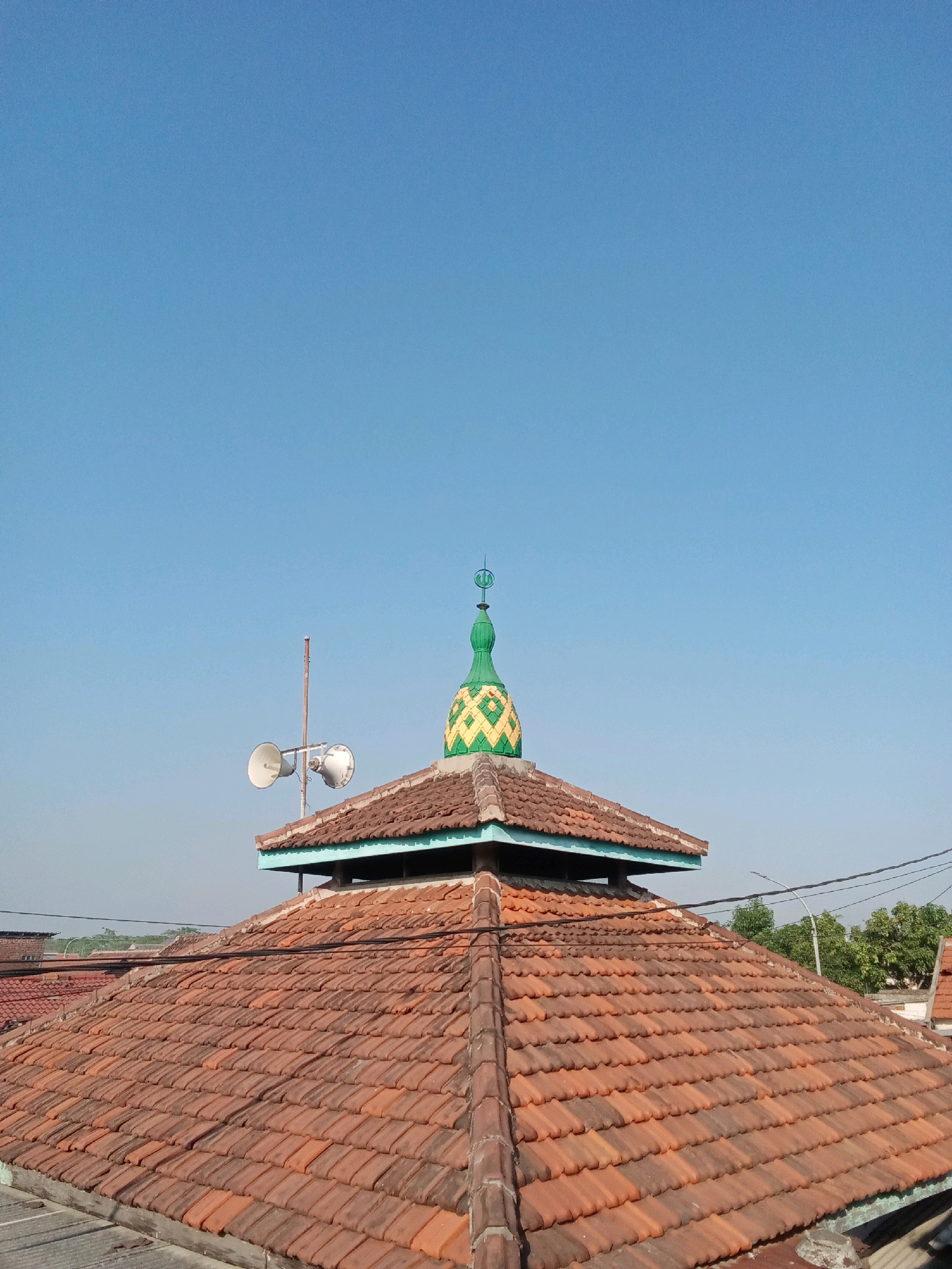 The image, taken from a rooftop perspective, captures a clear blue sky over the city of Sidorejo, Indonesia. In the foreground, a large, multi-faceted roof covered in terracotta tiles dominates the lower half of the frame. The tiles are laid in overlapping rows, displaying variations in color from orange to dark brown, suggesting age and exposure to the elements. A smaller, tiered roof structure is visible in the middle ground, topped with a vibrant green and yellow dome. This dome features a decorative pattern and is crowned with a finial. To the left of the dome, a tall pole stands, supporting two white loudspeakers. In the background, the hazy outlines of other buildings and lush green trees are discernible against the bright sky, hinting at a residential or mixed-use urban environment. The scene is devoid of any people or animals.