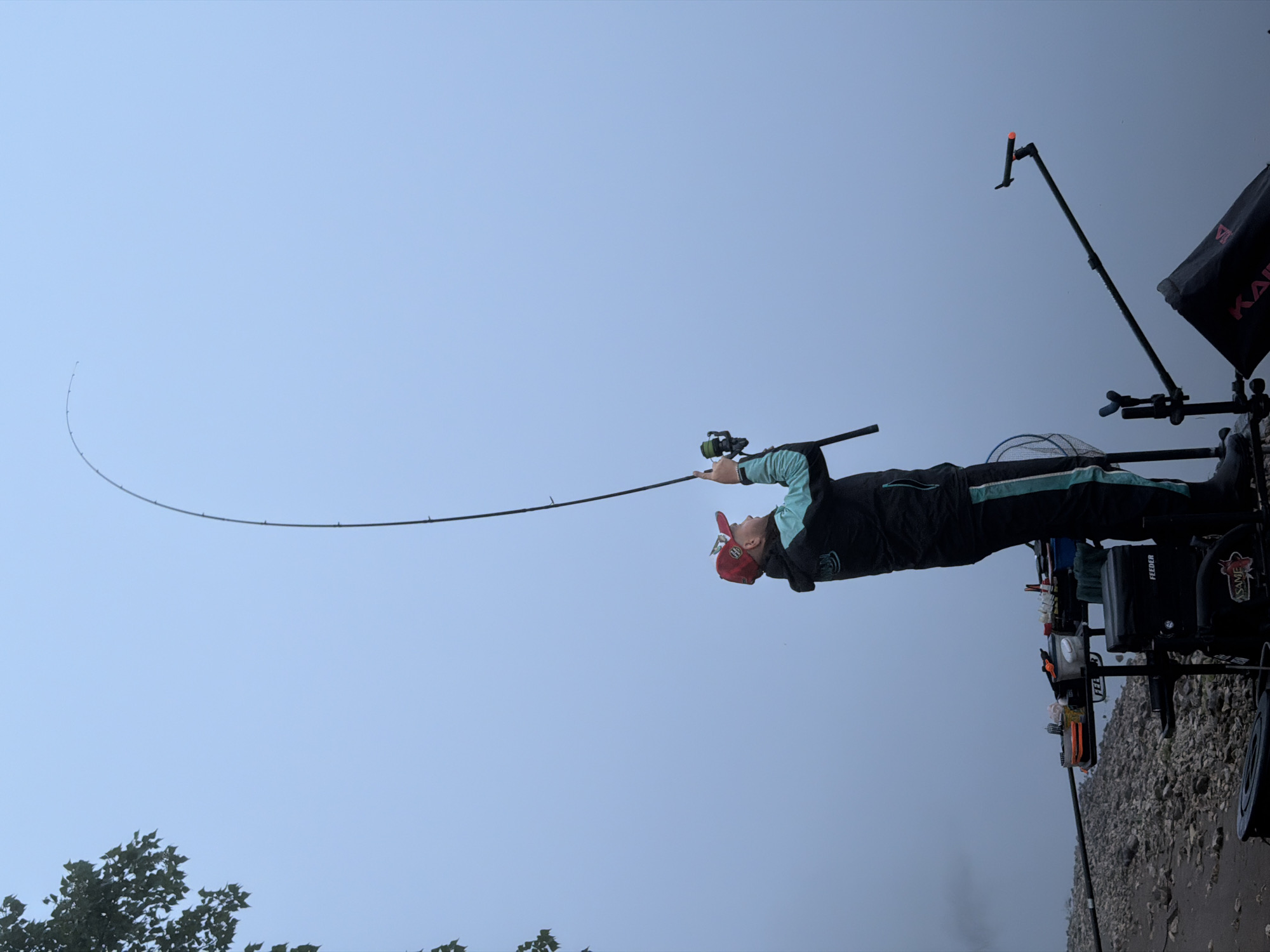 This image captures a person fishing on a rocky shore in Chaikovsky, Russia, under an overcast and misty sky. The angler, dressed in a black and teal jacket, black pants, and a red baseball cap, is intently focused on a long fishing rod that is significantly bent, suggesting a catch or strong tension. A fishing reel is visible on the rod.
Beside the angler is an elaborate fishing station, featuring a specialized seat or platform, multiple containers, and rod holders. A fishing net is also present among the gear. To the right, a black fabric canopy or tent displays the text "KAPR". Other visible text on the equipment includes "FEEDER" and "ASAME".
The setting is a natural outdoor environment, with a foreground of grey, uneven rocks or pebbles leading to what appears to be a large body of water, shrouded in haze. In the far background on the left, some tree foliage is faintly discernible. The sky is a uniform, light blue-grey, indicative of misty or cloudy conditions, and the diffused light suggests daytime. The overall atmosphere is one of serene concentration on the fishing activity.