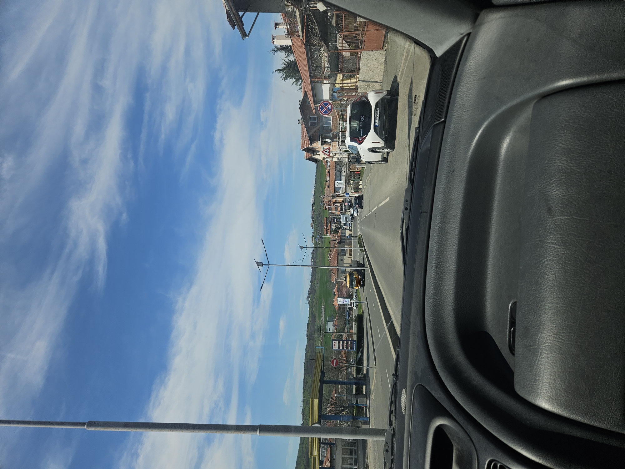 Captured from inside a vehicle, the image shows a street in Burgas, Bulgaria, on a bright, partly cloudy day. In the foreground, the car’s dashboard is visible, with the street extending into the distance through the windshield. The road has multiple lanes and seems to be relatively busy with traffic. Buildings, houses, and trees line the road, creating a typical suburban landscape. Streetlights are spaced along the road. At least one visible sign indicates "No Parking". The overall impression is of a common thoroughfare on a clear day.