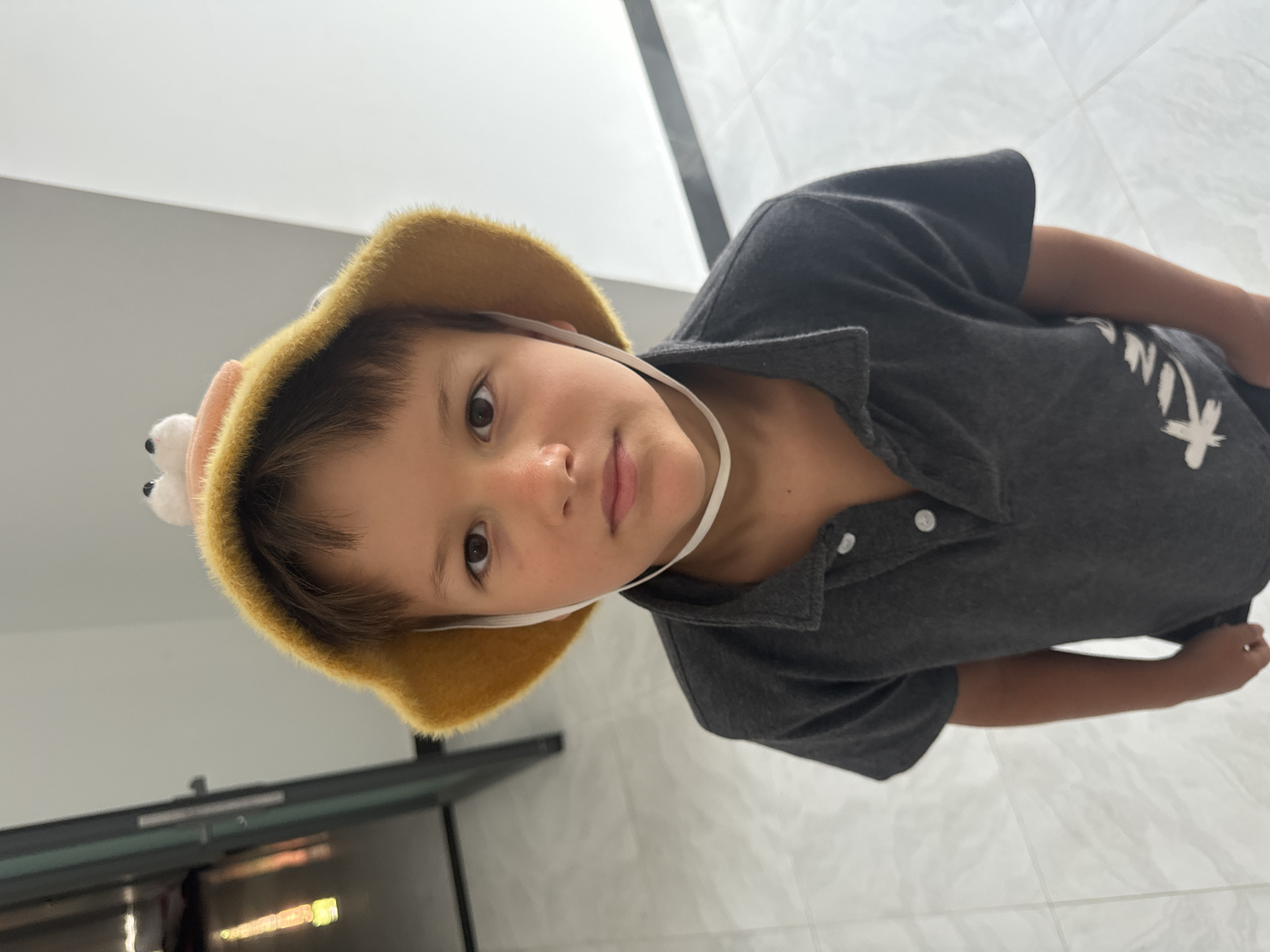 A young boy is indoors in Mũi Né, Vietnam, wearing a fuzzy tan hat with a white strap. He is wearing a gray collared shirt with the word "KIND" printed on it. The environment consists of white tiled floors and walls. There is a dark gray cabinet or object visible in the bottom left corner.