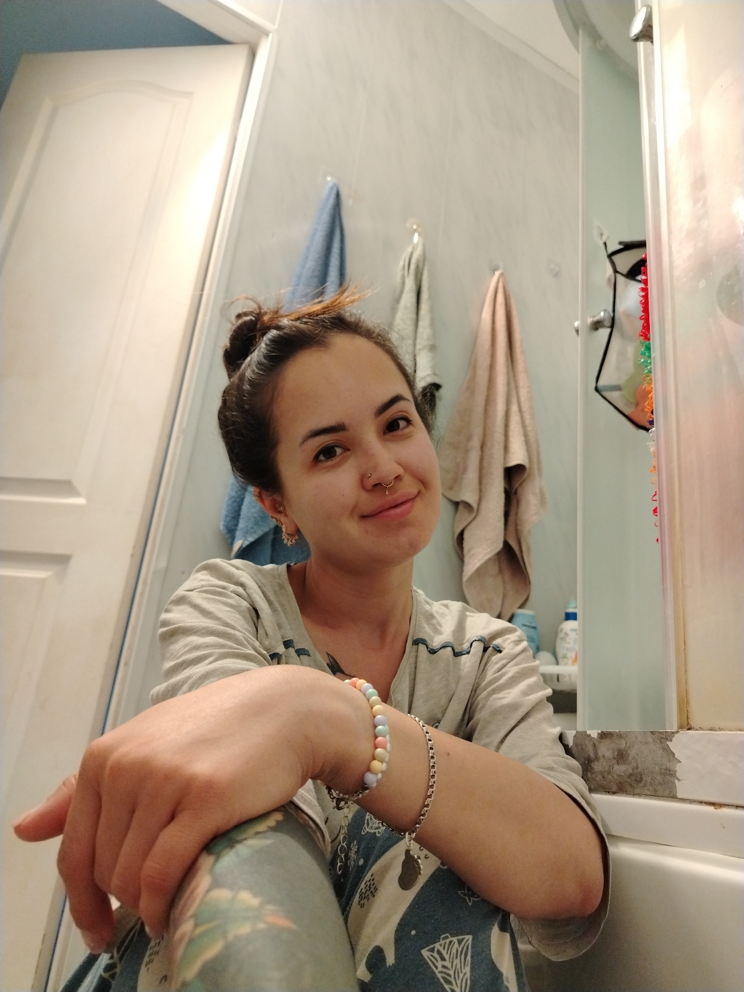 A young woman is featured in a low-angle indoor portrait, captured in what appears to be a bathroom in Noviy, Russia. She is seated, looking directly at the camera with a subtle, friendly smile and a calm expression. Her dark hair is styled in a messy bun atop her head. She is wearing a light-colored, long-sleeved top with a dark wavy pattern on the trim, and patterned pajama bottoms. On her left wrist, she sports two bracelets: one made of pastel-colored beads and a separate silver chain. She has multiple facial piercings, including a septum ring and a stud on her left nostril, along with small hoop earrings.
The background reveals typical bathroom elements. A white door is partially visible to the left. Behind the woman, blue and beige towels hang on hooks against a light-colored tiled wall. To her right, part of a shower or bathtub enclosure is visible, with a clear plastic caddy holding various toiletries, including bottles, hanging inside. The edge of the tub or shower base shows some signs of wear. The lighting is ambient indoor light. No text is visible in the image.