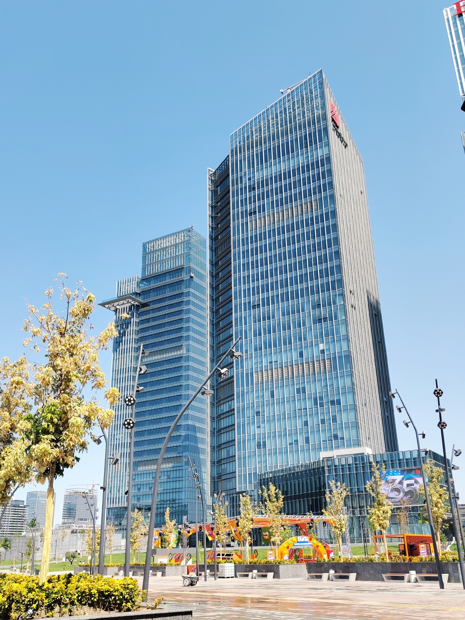 The image shows the Tashkent City International Business Center in Tashkent, Uzbekistan. Two modern skyscrapers, clad in blue glass, dominate the skyline. One of the buildings displays a red logo on its upper section. In the foreground, a yellow-leafed tree stands to the left, while to the right, there are streetlights and an outdoor event setup with colorful banners and seating. A small utility cart is visible near the event area. The sky is clear and blue, indicating a bright, sunny day. The scene appears to be a public plaza or park area adjacent to the business center.