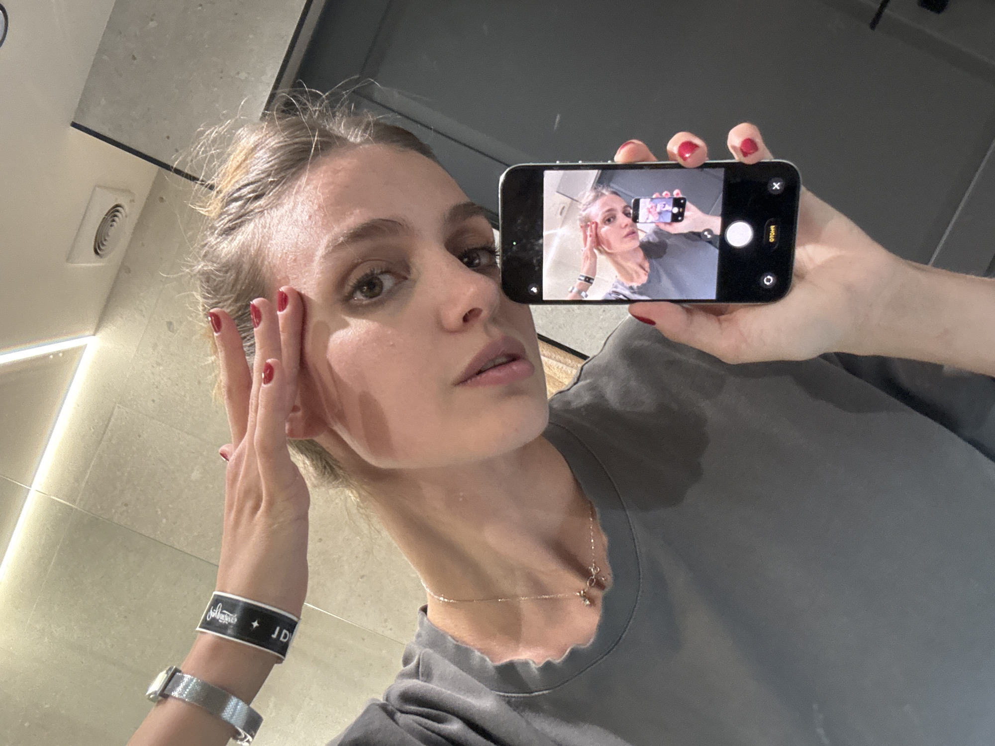 A woman with light skin and brown hair is holding a smartphone in her right hand, taking a picture of herself. The phone displays an image of herself taking the picture. She is wearing a gray t-shirt and a delicate gold necklace with a pendant. On her left wrist, she has a silver watch and a black wristband with white writing. Her fingernails are painted red. She is standing in what appears to be a bathroom, with light-colored tiles and a ventilation fan visible on the wall. The lighting is bright and even. There is no indication that this photograph was taken in Yerevan, Armenia.