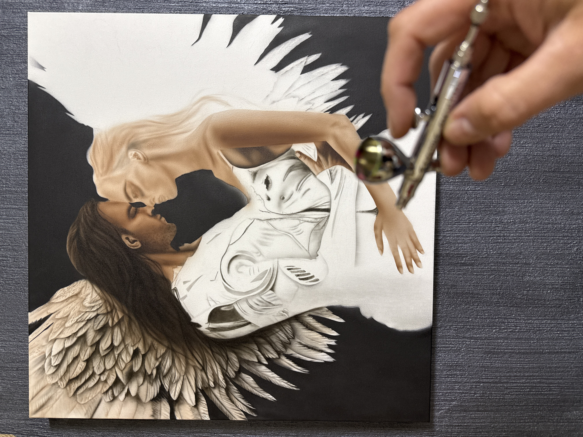 The image shows a painting of a man and woman with wings embracing. The woman has blonde hair and light skin, while the man has dark hair and darker skin. They are set against a black background, with the wings appearing in shades of white and gray. The painting appears unfinished as large portions of the canvas are still white. A hand holding an airbrush is visible in the upper right corner of the image, suggesting the painting is in progress. The background texture on the table is a dark gray and looks to be a rough material. There is no indication of a city or location in Burgas, Bulgaria.