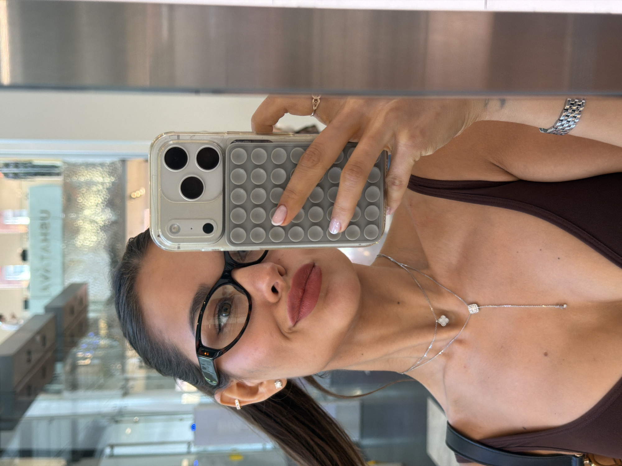 An image, captured as a mirror selfie, is rotated 90 degrees clockwise, showing a young woman looking into the camera. She has dark hair, styled away from her face, and wears black-framed glasses. Her lips are a muted reddish-brown. She is adorned with several pieces of jewelry: small stud and hoop earrings, a silver-toned multi-layered necklace featuring a small, possibly four-leaf clover-shaped pendant and a lariat-style chain with a square, sparkling element, and a metallic, woven-patterned watch on her left wrist. A small ring is also visible on her right hand.
The woman is holding an iPhone, likely a Pro Max model given the prominent camera array, encased in a clear cover with a gray "pop-it" style panel on the back. Her nails are manicured; one hand shows a classic French tip, while the other features a pearlescent finish. She wears a dark brown or maroon-colored top with thin straps.
The background, a reflection in the mirror, suggests an indoor, modern setting, possibly a public space like a shopping mall or a building lobby in Moscow, Russia. Blurred architectural elements, such as light-colored walls, metal structures, and distant lights, are visible. Reflected text, reversed, can be partially discerned as "УАТАНУА" (likely "AVATANU" or similar in Cyrillic), further supporting the location context. The overall lighting is bright and even, indicating a daytime or well-lit interior environment. The woman's expression is neutral as she captures her reflection.