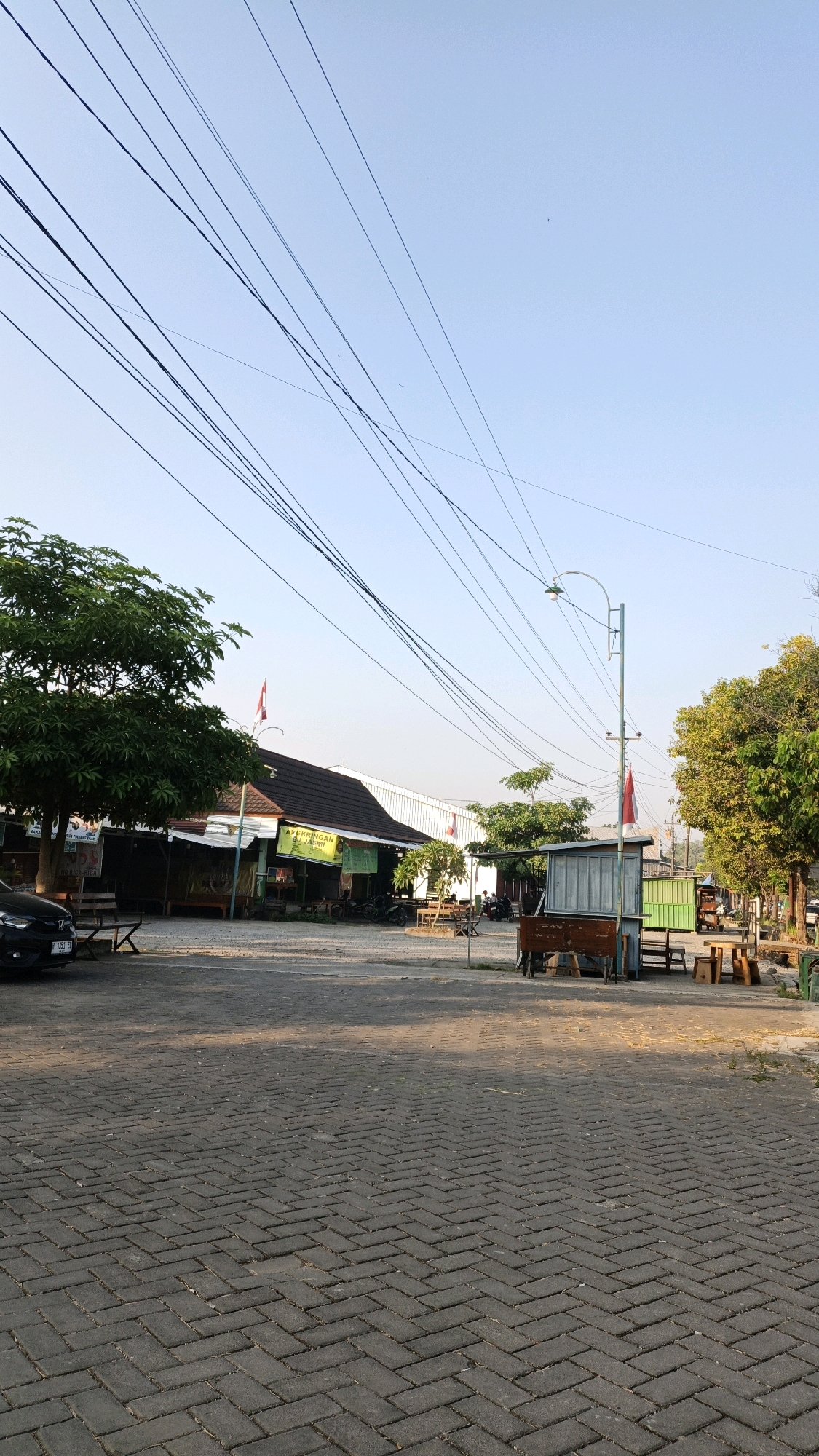 The image depicts an outdoor scene on a clear day, likely in Kuwasen, Indonesia. The foreground is dominated by a paved area with interlocking grey brickwork laid in a herringbone pattern. In the mid-ground, there are several structures and elements typical of a roadside establishment or market. A building with a dark tiled roof and white walls is visible on the left, with a green sign that reads "NASI CIKRING & RUJAK JARMI." A large leafy tree stands to the left of this building, and a black car is parked in front of it. To the right of the building, there are various wooden tables and benches, suggesting a seating area. Further to the right, a small open-air stall with a grey corrugated metal roof and wooden front panels is situated near a light pole. Beyond this, more seating and structures, including a green container-like building, are visible. Multiple power lines crisscross the sky, which is a pale blue. The overall impression is of a casual, possibly a food court or a cluster of small eateries, operating in daylight. There are no people prominently featured, though some activity might be inferred by the presence of the parked vehicles and the ready-to-use seating.