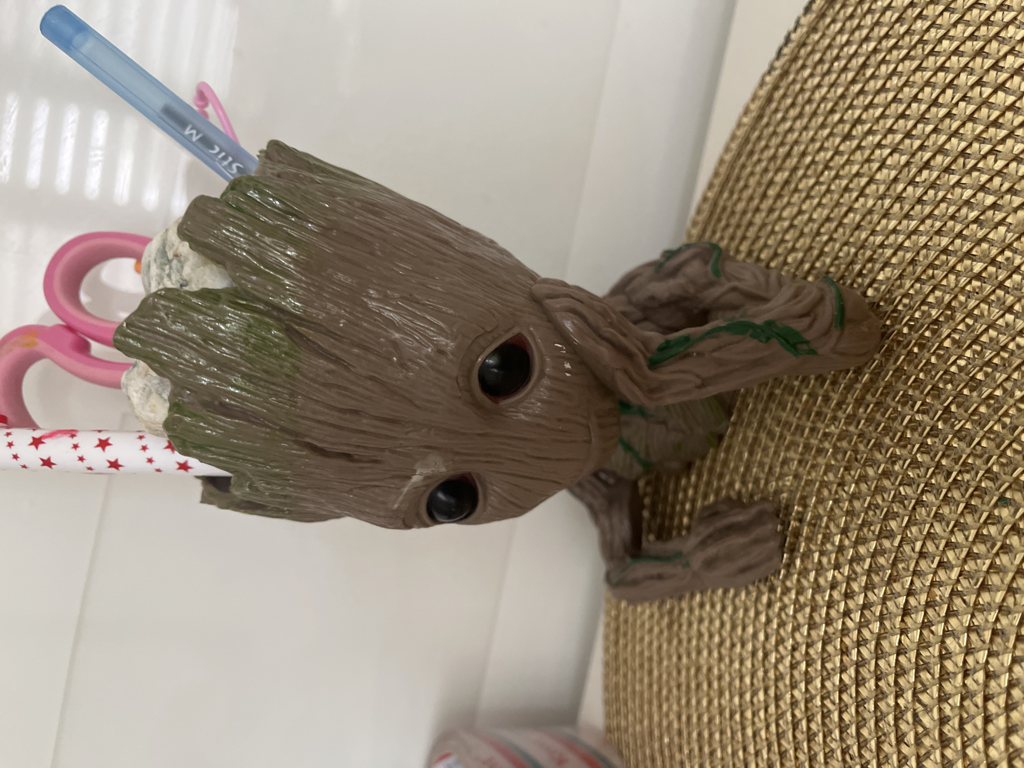 An indoor, close-up photograph displays a brown, textured plastic figurine resembling the character Groot, acting as a pen holder. The figurine is positioned in a pensive pose, with its left hand supporting its chin and its right arm resting forward. Green accents, simulating foliage, are present on its body. Inside the hollowed-out top of the figure, a white pen adorned with red stars, a translucent light-blue ballpoint pen marked "Stic M," and the pink handle of a pair of scissors are visible. Small, irregular white objects are also present within the holder. The Groot figurine rests on a gold-colored, tightly woven mat with a distinct geometric pattern. The background consists of a plain white wall or surface. The location is an unspecified indoor setting in Unknown, Unknown.