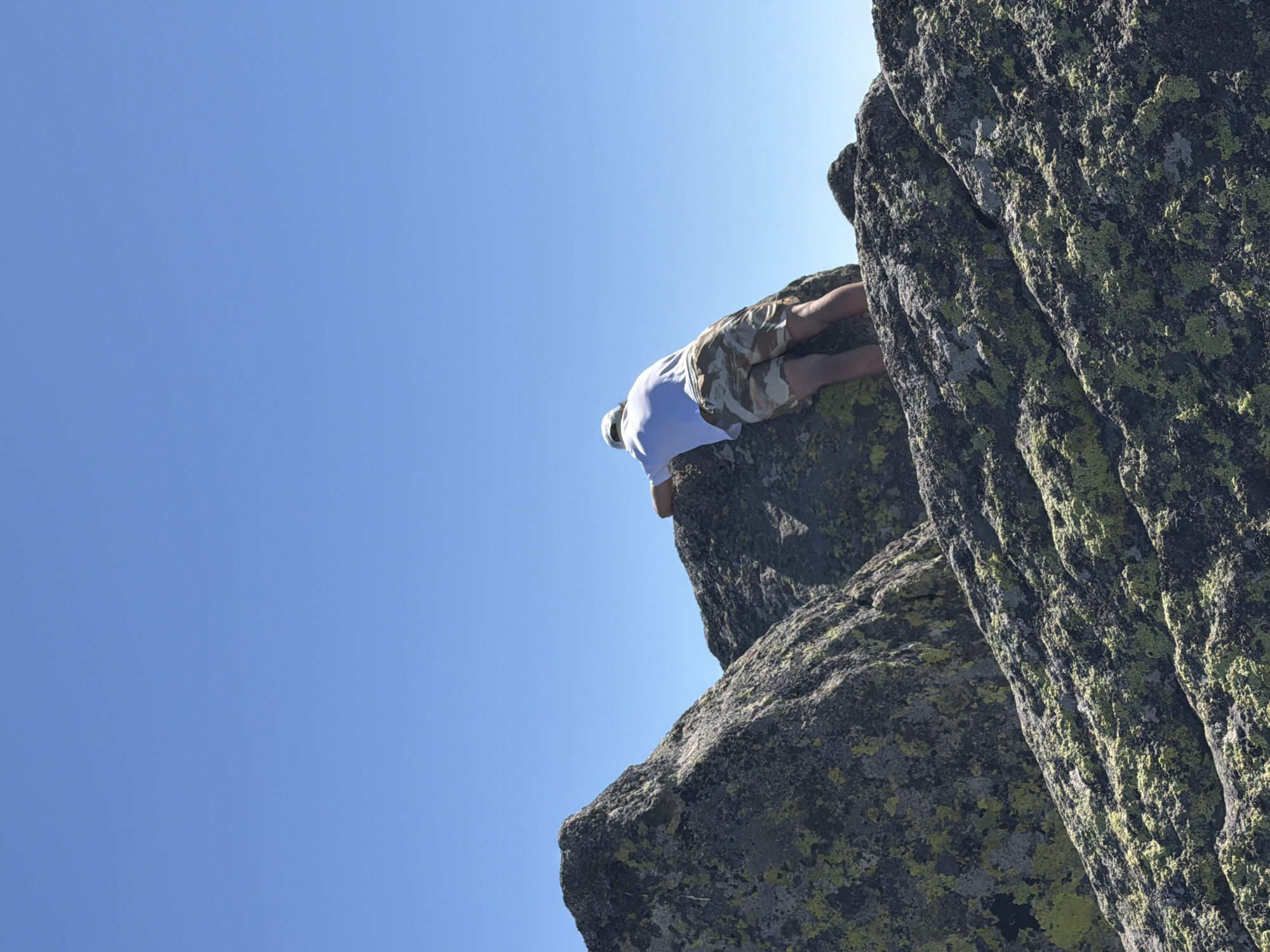 A person, seen from behind, is leaning over large rocks under a clear blue sky. The individual is wearing a white t-shirt and camouflage shorts, with a light-colored cap on their head. They appear to be standing on a rocky mountain surface, possibly Vitosha Mountain, near Bistritsa, Bulgaria. The rocks are covered in patches of green and yellow lichen. The person's hands are resting on the rock in front of them.