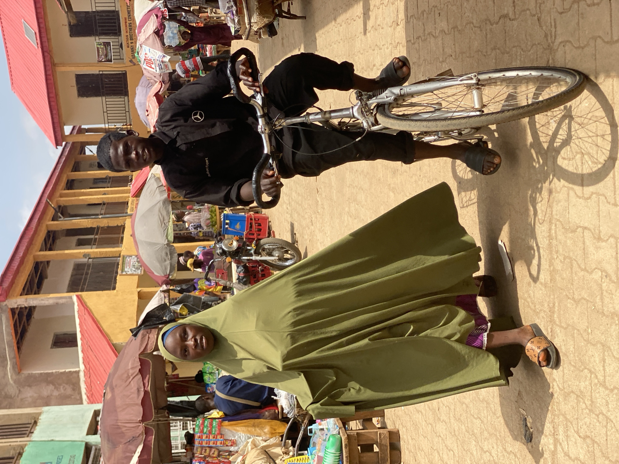 A photo depicts two individuals in a bustling street scene in Kafanchan B, Nigeria. A young man stands beside a silver bicycle, wearing a black shirt, black pants, and black sandals. He grips the bicycle handles. Beside him, a woman stands cloaked in a green garment, and a portion of her patterned purple and white garment is visible.
The street is paved with patterned tiles. Umbrellas shade vendor stalls. Other people, motorcycles, and merchandise fill the background. Buildings with red roofs and yellow walls flank the scene.