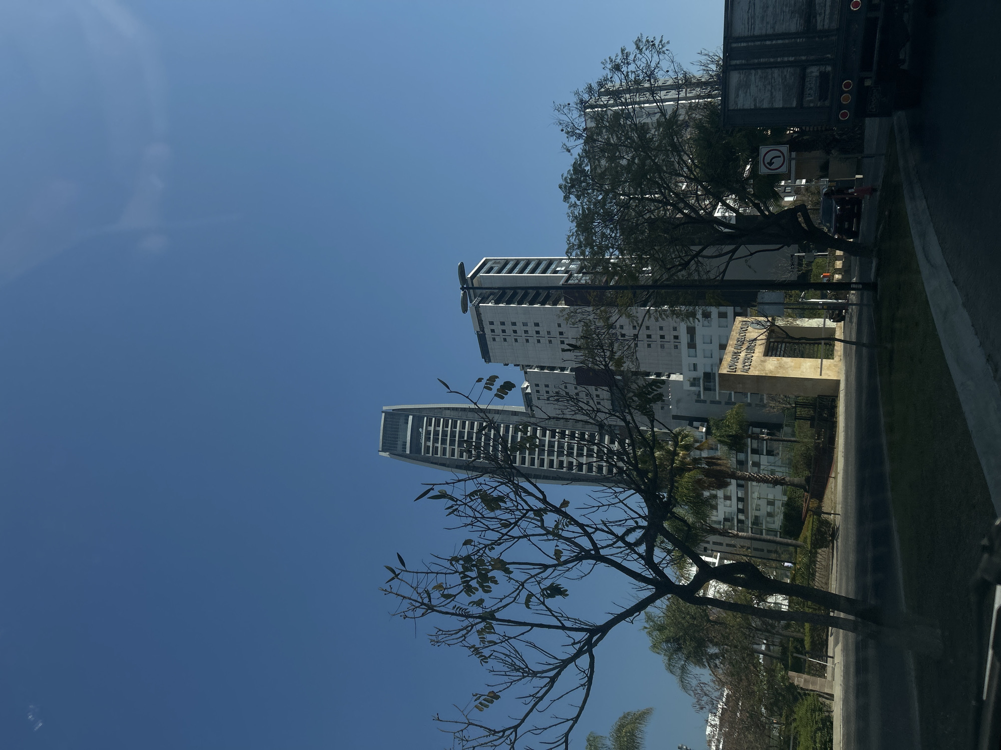 The image depicts a street scene in San Bernardino Tlaxcalancingo, Mexico. Two tall, modern buildings with a light facade are visible in the background. Bare trees frame the buildings. A large truck is parked along the road with a no turn left sign, casting a shadow on the street. A dark-colored car is partially visible behind a tree. A sign at the entrance reads "LOMAS DE ANGELÓPOLIS ACCESO VECINOS." The sky is clear and blue.