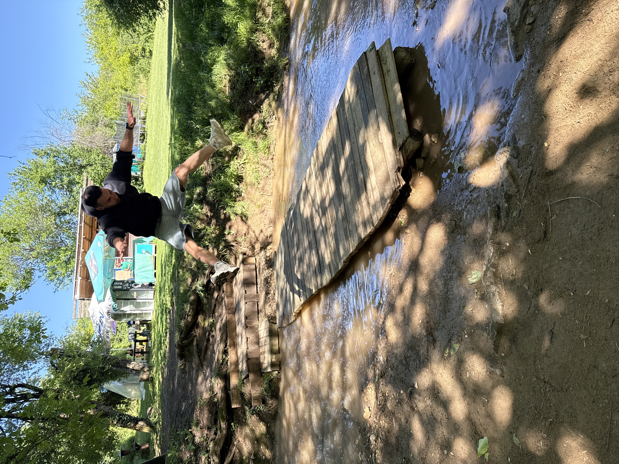 A man in a black shirt and gray shorts is jumping over a water-filled ditch spanned by a wooden bridge. He is mid-jump, with one foot on a makeshift wooden staircase and the other extended in the air. The scene is outdoors, with grassy areas, trees, and structures in the background. The location is Hotnitsa, Bulgaria. The water in the ditch is murky. The ground is muddy and uneven.