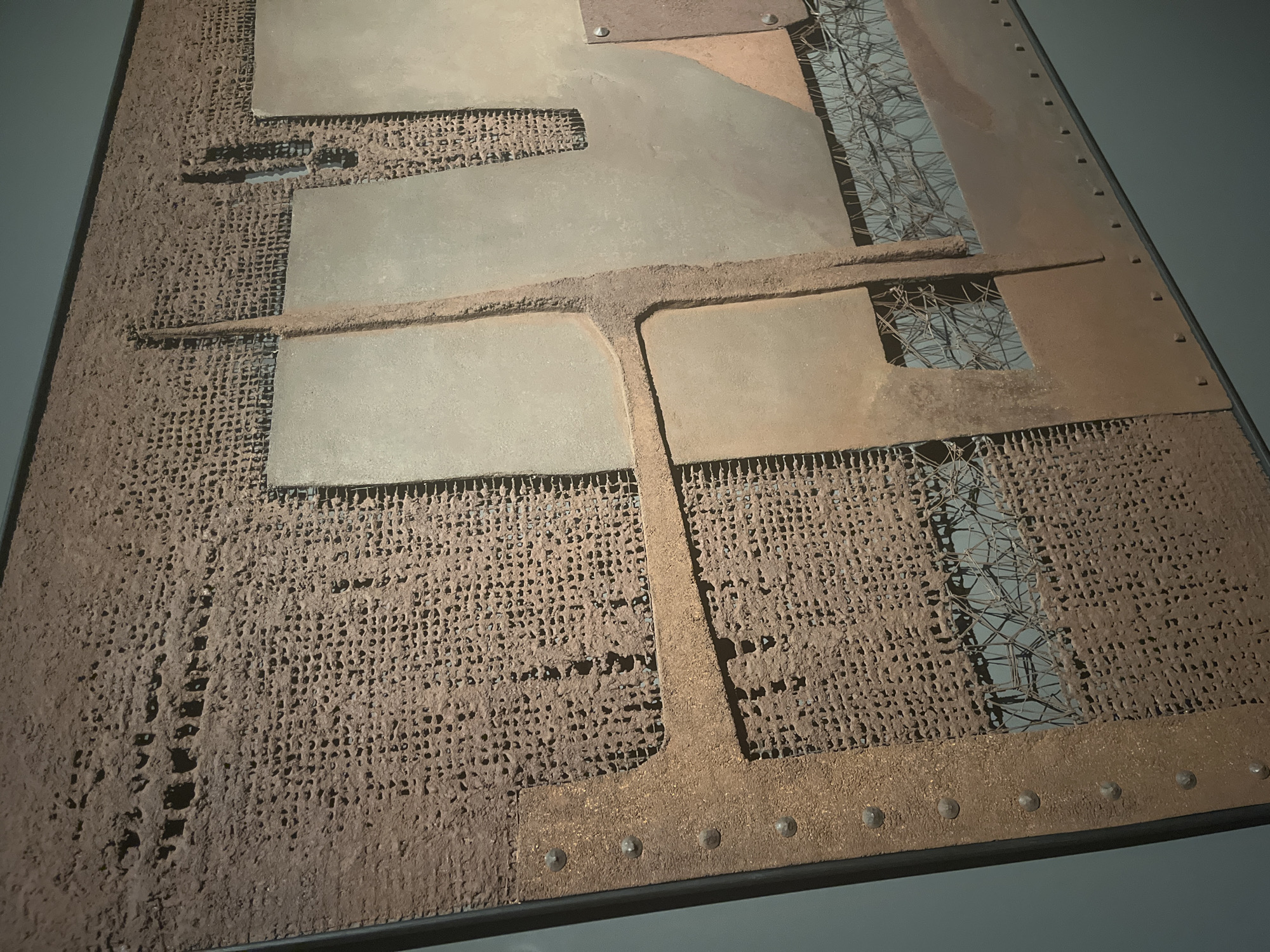 This image captures a close-up of a textured, mixed-media artwork, likely displayed in a museum or gallery setting in València, Spain. The artwork is characterized by its earthy, industrial aesthetic and layered construction, devoid of people or activity.
The piece features a rectangular composition built upon a dark, possibly metallic frame visible along its edges. The primary surface is covered with a reddish-brown, porous, granular material, resembling sand or heavily corroded earth, applied over a fine mesh or netting which is visible in areas where the granular layer is thinner.
Overlaid on this textured base are several flat, rectangular and irregularly shaped panels. Some panels are a muted, light grey, appearing metallic and relatively smooth, while others are a darker, rusty brown, suggesting oxidized metal. These panels are arranged in a grid-like but staggered fashion. A prominent, raised T-shaped strip, made of the same granular material, traverses the surface, separating some of the underlying textures and panels.
Through rectangular cut-outs in some of the panels and the granular layer, a coarser, metallic wire mesh with a wider, more open pattern is visible, adding depth and another layer of material. The right edge of the artwork, and some other sections, are adorned with a row of small, rounded protrusions, likely rivets or fasteners, enhancing its industrial character. The overall color palette is dominated by various shades of brown, grey, and rust, creating a weathered and robust impression. No text is visible on the artwork.