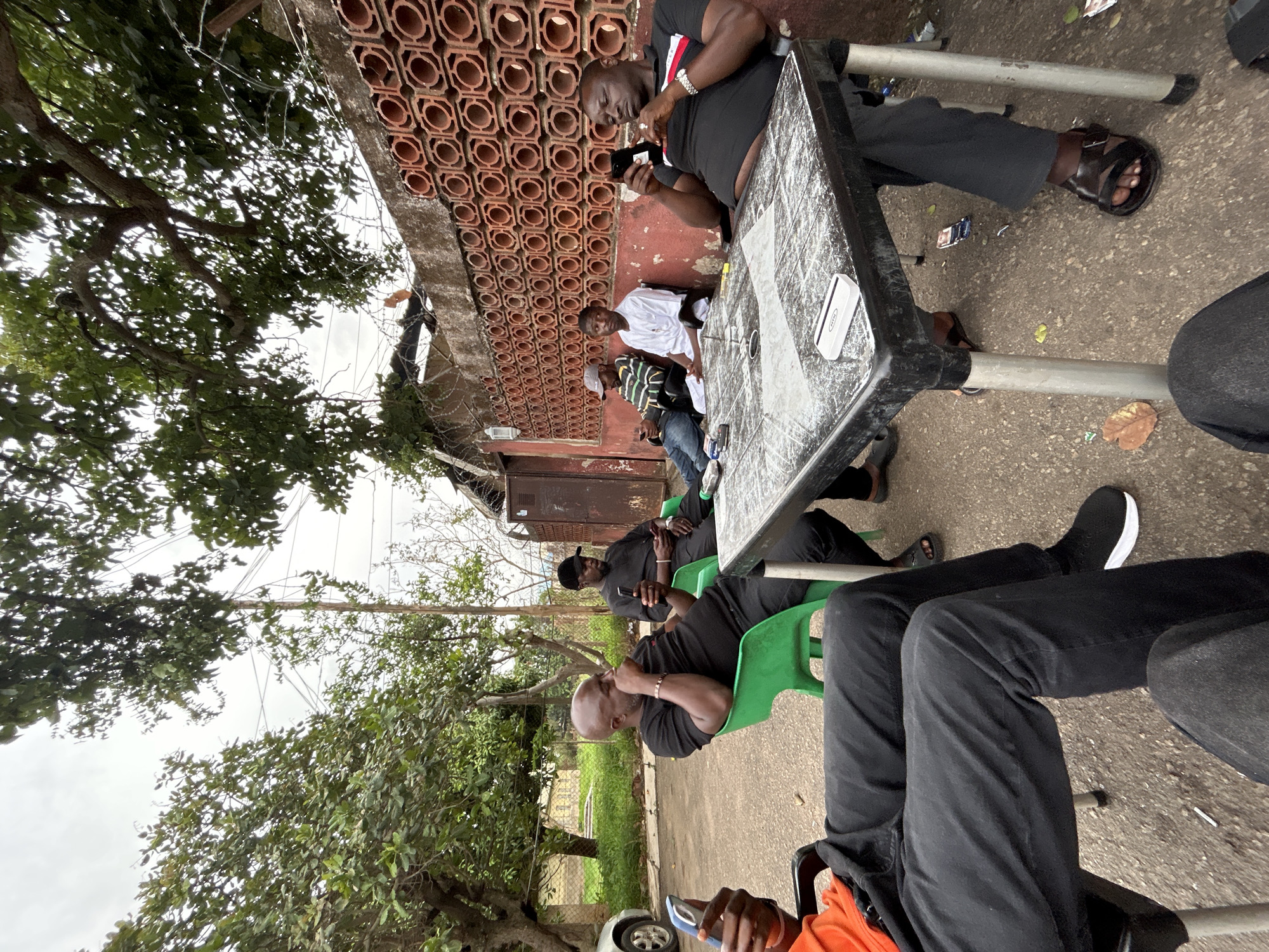A group of men are sitting at a table outdoors in Jos, Nigeria. Most of the men are looking at their phones. There is a red brick wall in the background with decorative round holes. The area is shaded by trees, and there are power lines visible above. The table is black plastic, and the chairs are plastic. Some litter is visible on the ground.
