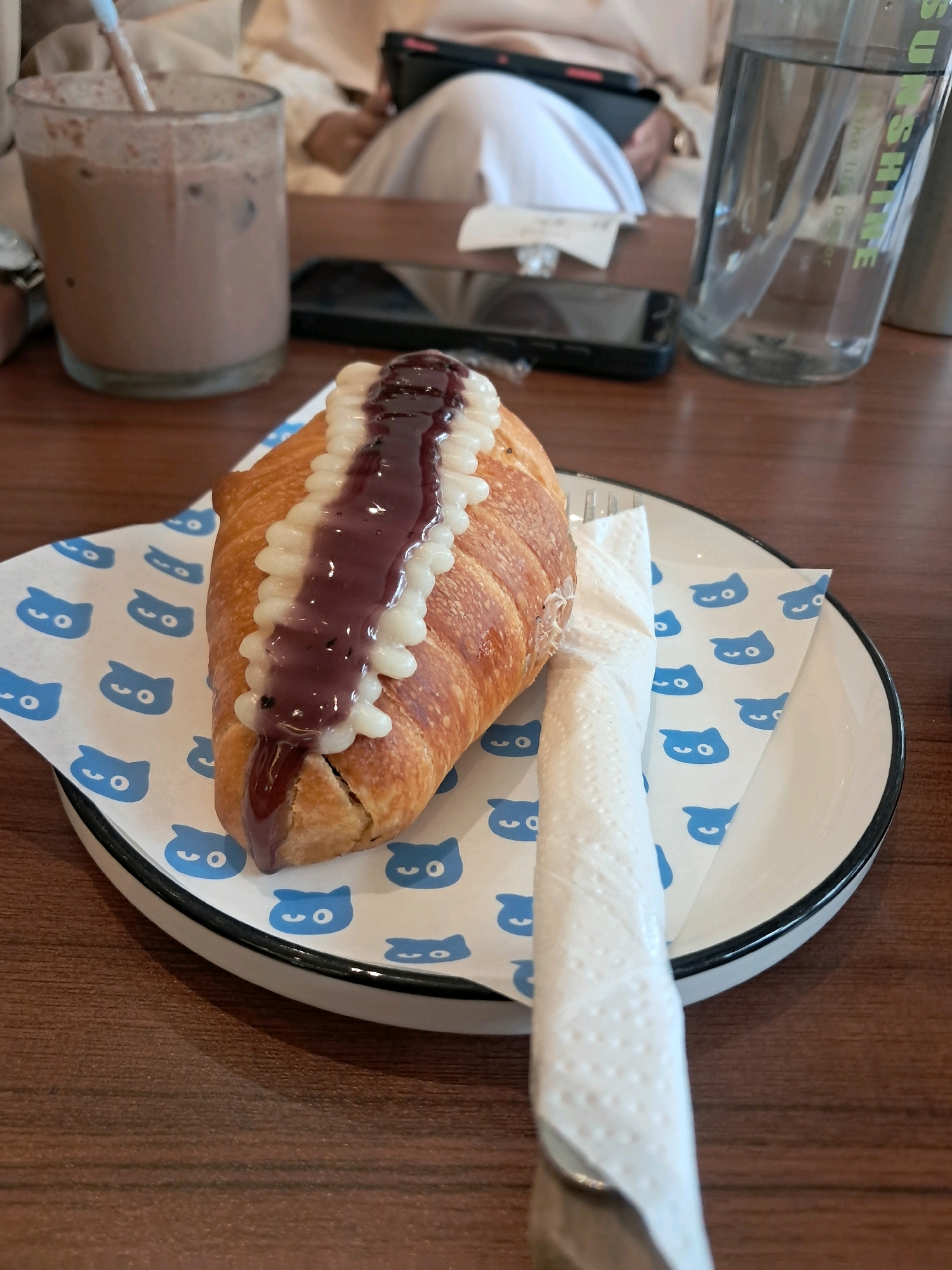 A close-up shot shows a croissant topped with white cream and dark jam on a plate with a blue cat pattern, resting on a wooden table. To the left, a glass of chocolate drink with a straw is visible. To the right, a clear bottle with a straw contains water, and a cell phone lies on the table. In the background, blurry figures of people are seated, suggesting an indoor setting like a cafe. The scene does not contain any specific visual elements that would allow for precise geolocation to Singkawang, Indonesia.