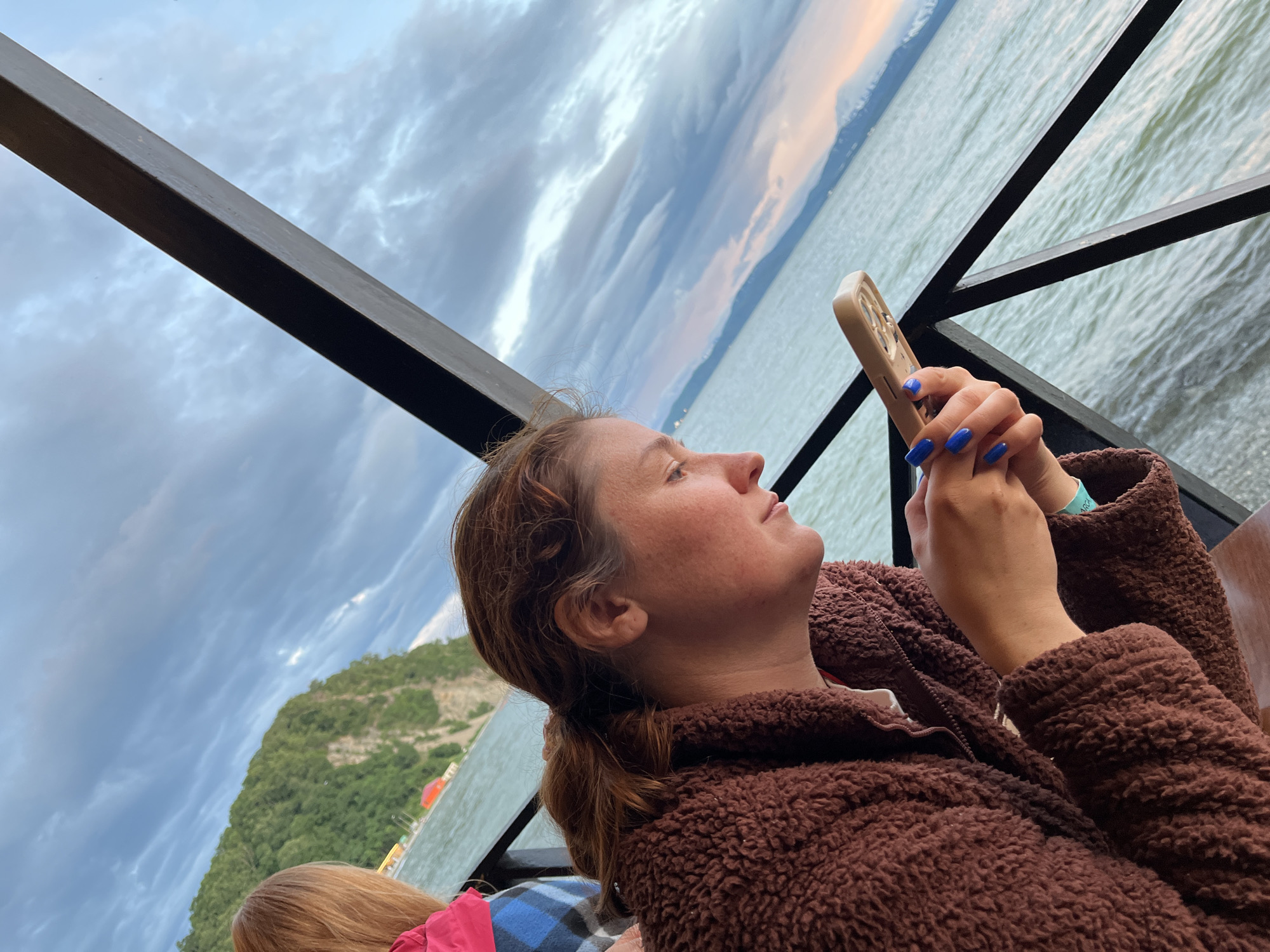 An outdoor shot captures a woman with auburn hair and freckles looking at her phone. She is wearing a brown fuzzy jacket. To her left, another person's blonde hair is visible, and they are wearing a pink and blue plaid shirt. The background shows a body of water, a steep, green, treed hillside, and a dramatic sky with clouds. A dark metal railing runs across the foreground. The setting appears to be a cafe or restaurant with outdoor seating. The overall mood is relaxed and informal.