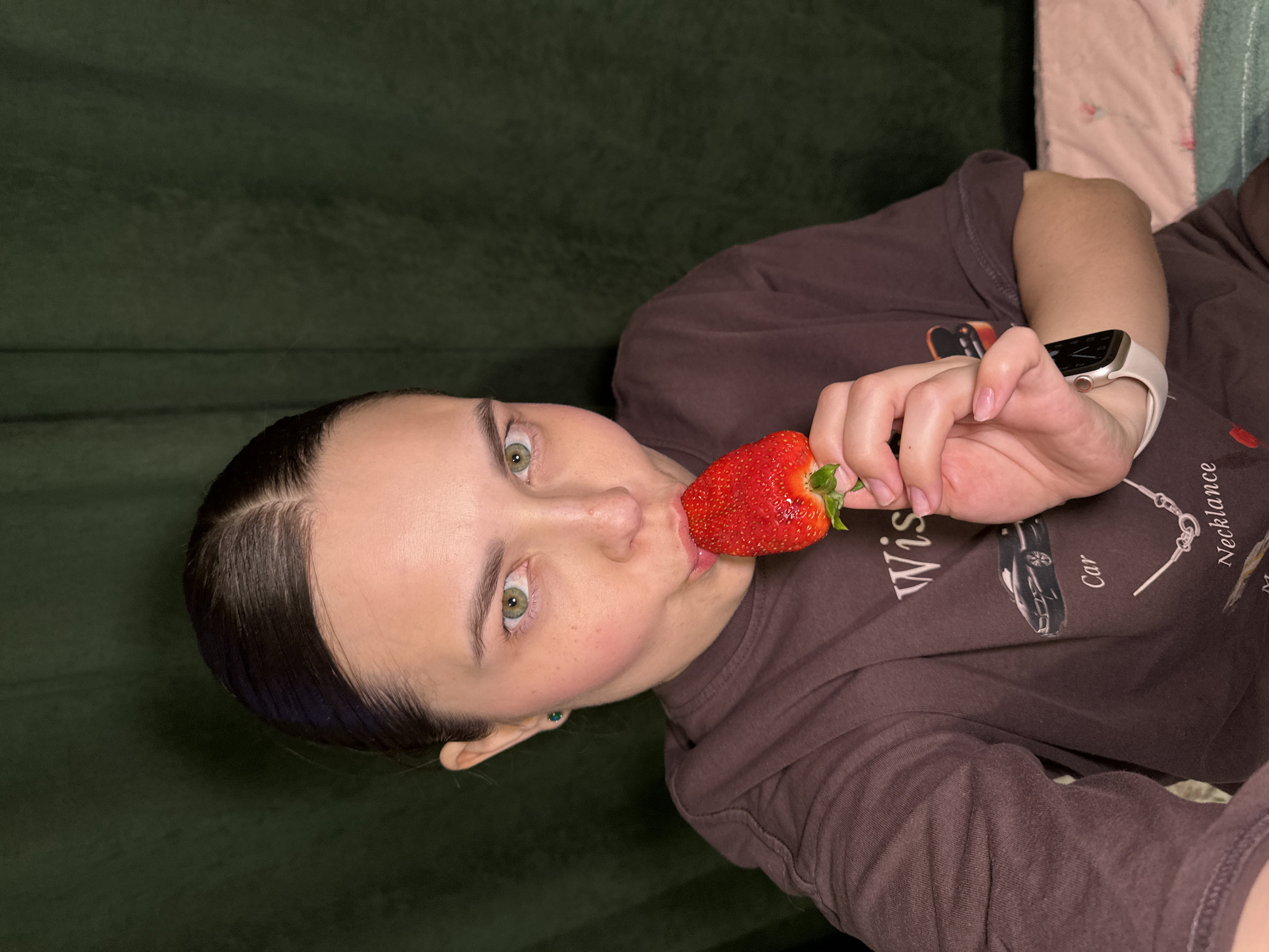 A woman is holding a large strawberry to her lips. She has fair skin, dark hair pulled back, and green eyes. She is wearing a dark brown t-shirt with graphics printed on the front, and an Apple Watch on her wrist. The background includes a dark green textured wall and a pink and green patterned blanket. The location, Perm, Russia, is not discernible from the image.