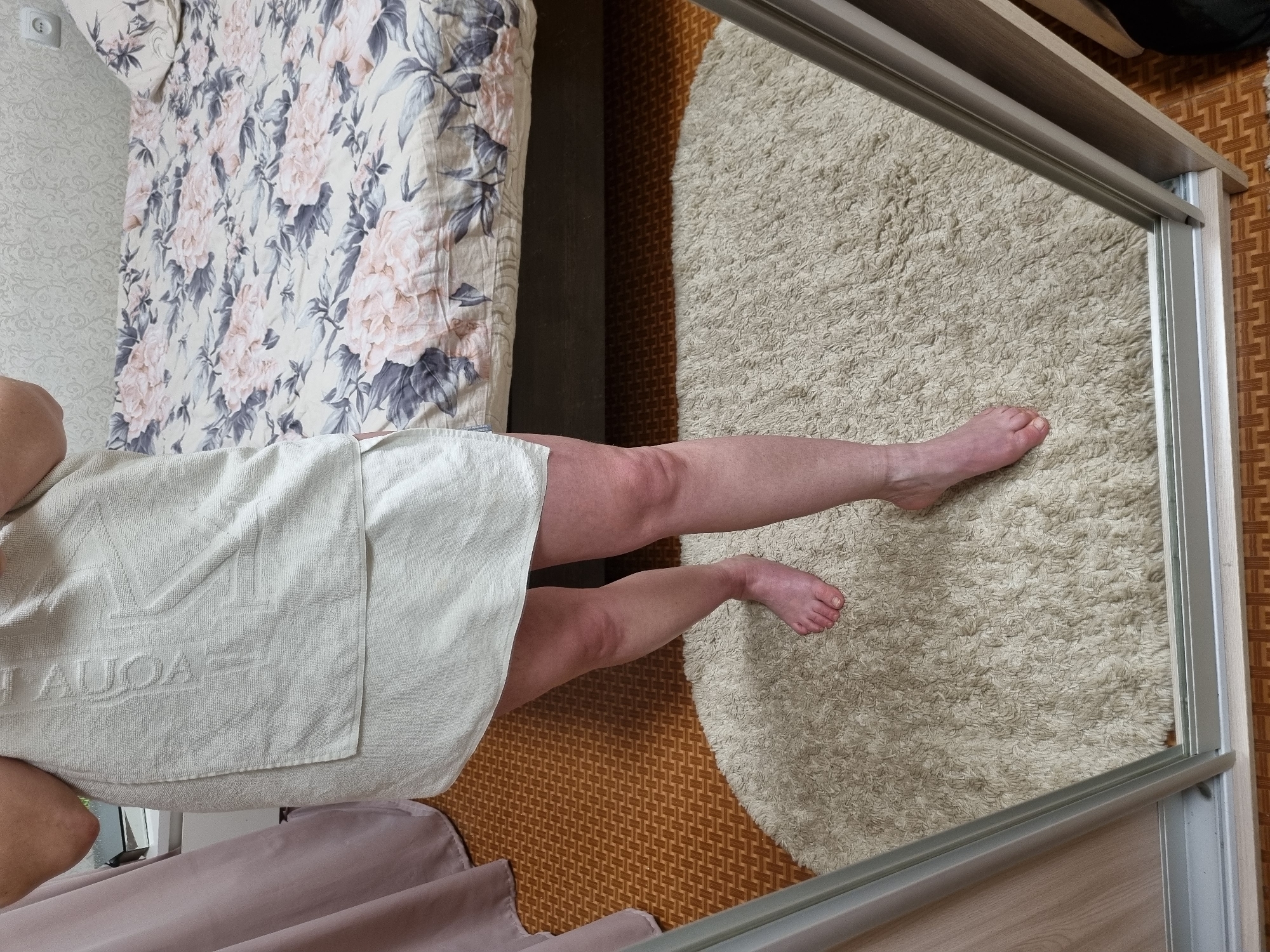 An overhead indoor shot captures a person wrapped in a light-colored towel, standing on a patterned floor in what appears to be a bedroom or dressing area in Chaikovsky, Russia. The individual's bare legs and feet are visible, extending into the frame from the left and center. The skin on the legs appears somewhat reddened. The towel has a textured surface with the visible text "MAUOA" embossed on it.
To the left of the person is a bed covered with a light-toned duvet featuring a large floral pattern of pink and grey blossoms and leaves. The floor is composed of two distinct sections: a fluffy, light beige rug partially reflected in a large mirror, and a brown or orange patterned mat. The mirrored surface, likely part of a wardrobe door or full-length mirror, reflects the rug and the person's lower legs and feet. A wall with subtly patterned wallpaper and an electrical outlet is visible in the upper left corner near the bed. The overall lighting suggests it is daytime. The image's perspective is rotated counter-clockwise.