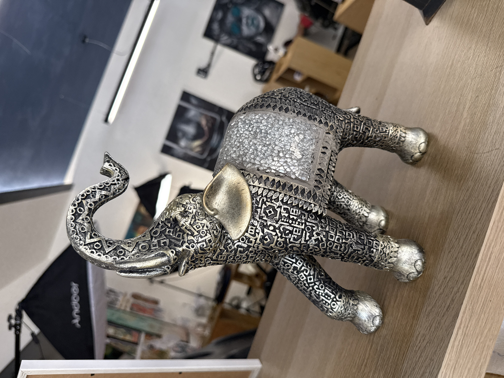 A silver elephant statue with intricate patterns is resting on a wooden surface. The statue has a decorative back with a mosaic-like texture. In the blurred background, there are art pieces on a wall, a softbox lighting equipment, and other furniture. There is not enough information to ascertain the location in the city of Sofia, Bulgaria.