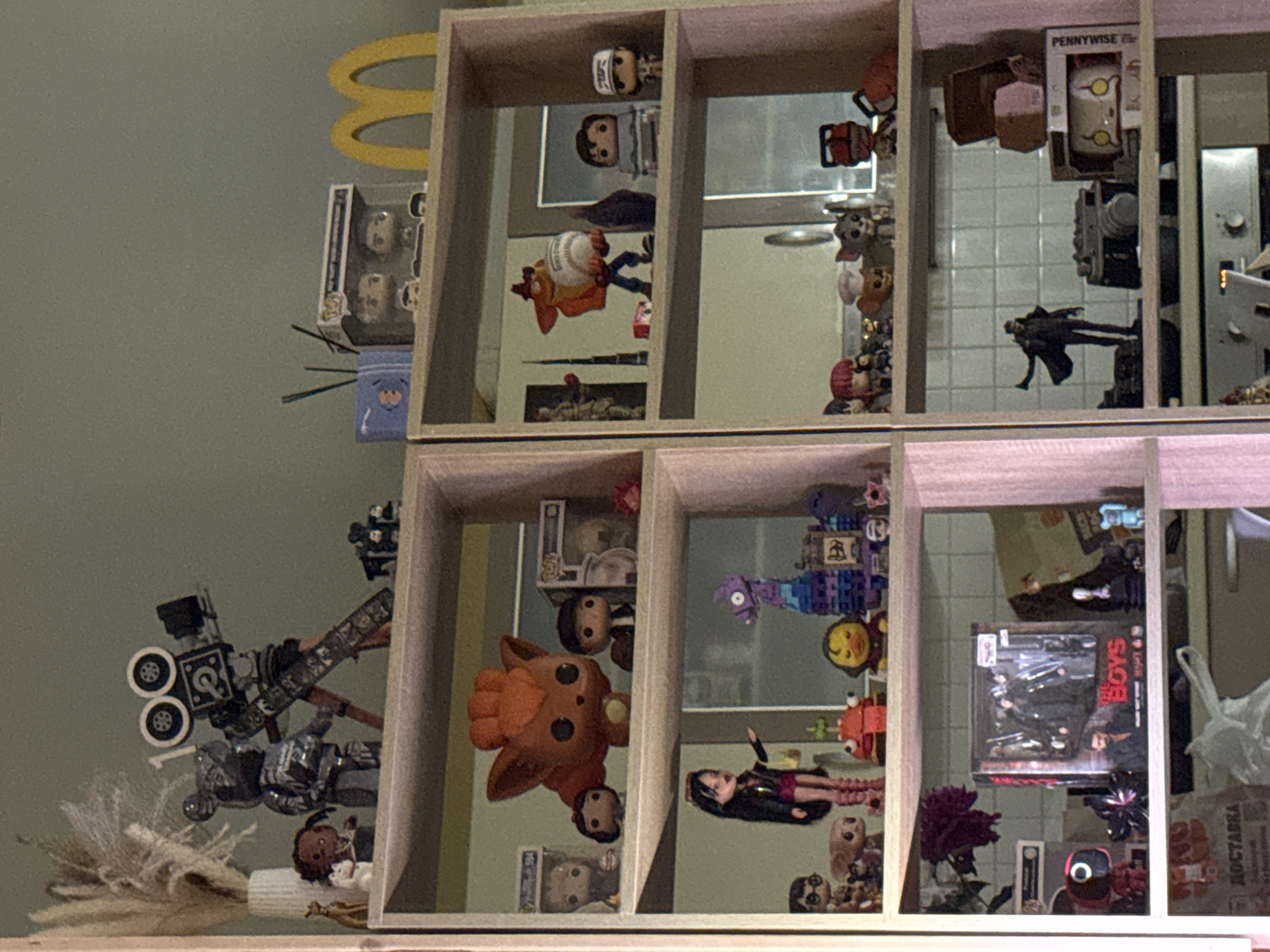 The image depicts a shelf filled with various collectible figures and decorative items. On the top shelf of the left side, there is a vintage film camera, a bear-shaped figure, and a vase with dried reeds. On the top shelf of the right side, there are two Funko Pop figures in boxes, a blue dispenser with dark sticks, and a yellow McDonald's logo. The middle shelf on the left holds a large orange creature figure, several smaller figures, and a doll. The middle shelf on the right displays a Crash Bandicoot figure holding a baseball, a figure of what appears to be a character from a game, and two more Funko Pop figures. The bottom shelf on the left contains a red Squid Game figure, a box set of "The Boys" figures, and other small figures. The bottom shelf on the right features a figure of a person in a long coat, a camera-like object, and a box with a Pennywise figure. The shelves are made of light-colored wood. The background appears to be a kitchen with tiled walls. The overall scene is an indoor display of pop culture memorabilia. The mention of Khimki, Russia, cannot be verified from the provided image.